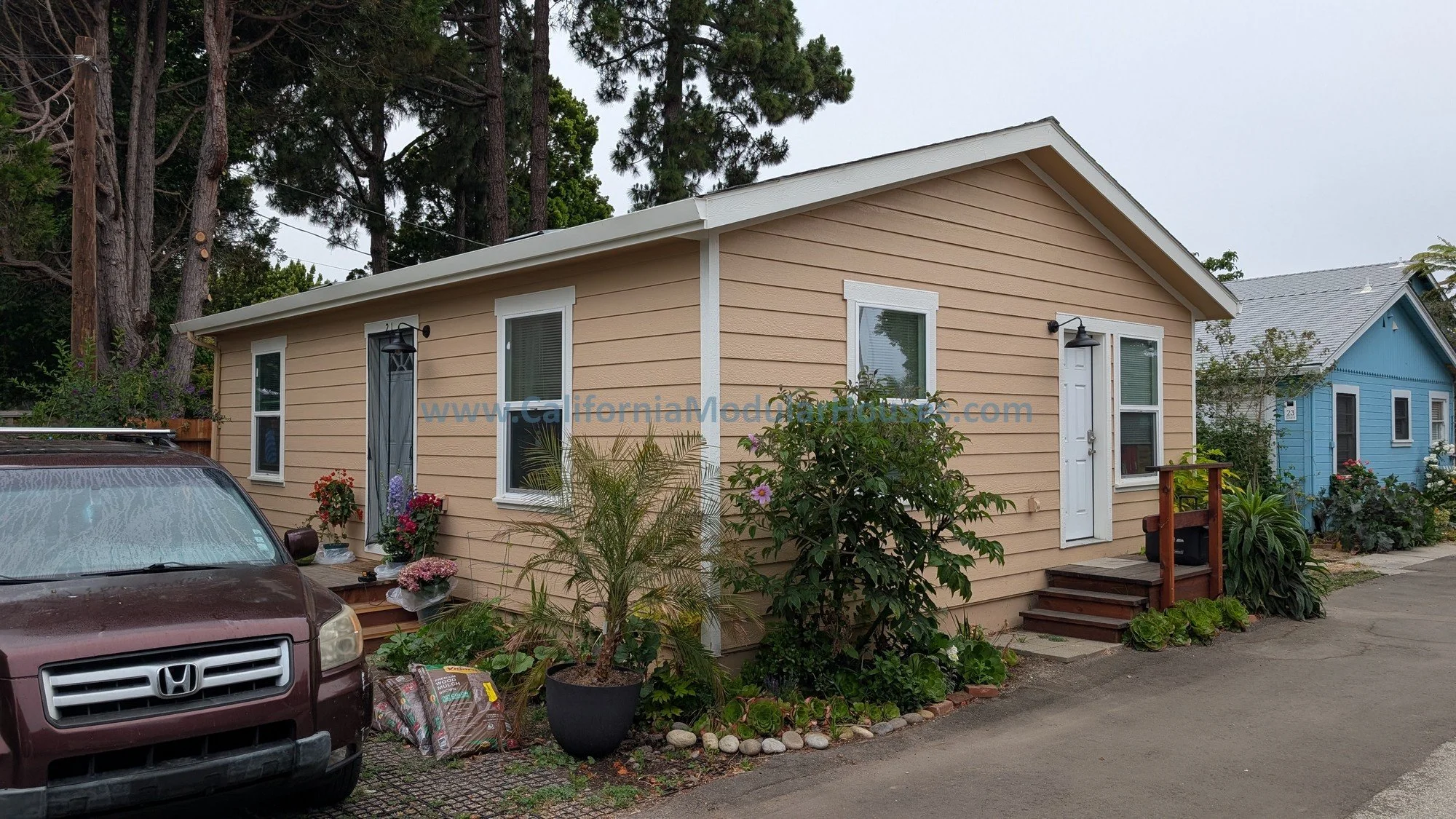 A beige single-story house with white trim and a small wooden staircase at the front door, surrounded by plants and flowers, with a maroon SUV parked in front on a paved driveway. There are trees in the background and neighboring houses painted blue.