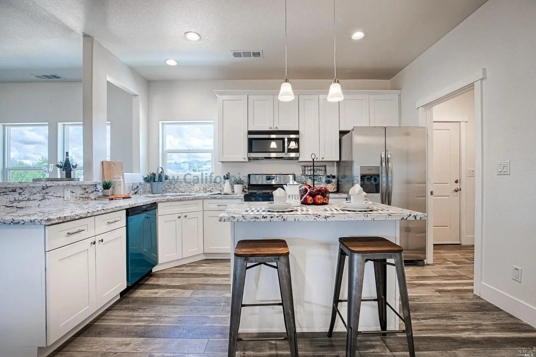 Modern kitchen with white cabinets, stainless steel appliances, granite countertops, a small kitchen island with two stools, and a window showing a partly cloudy sky.
