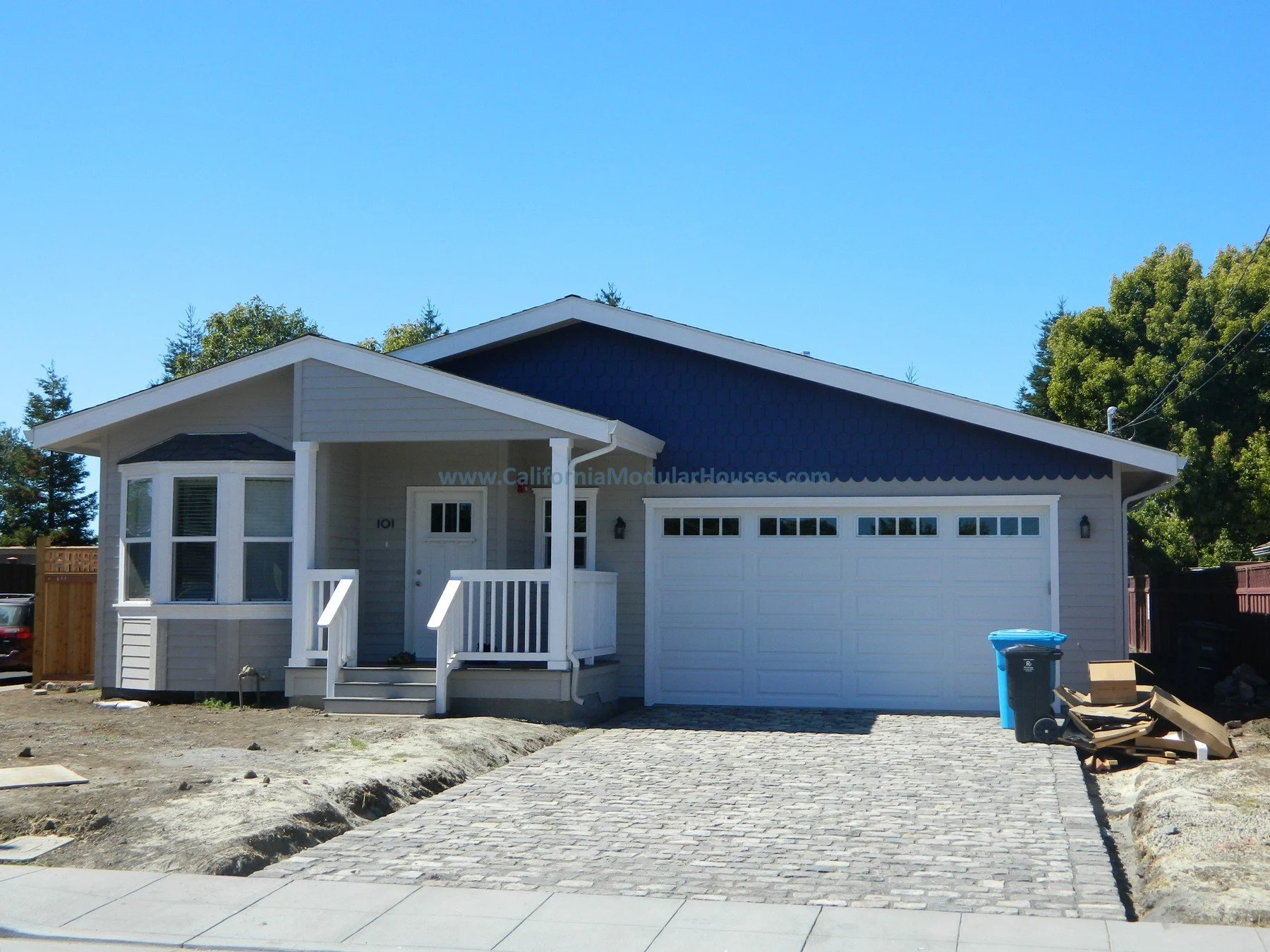 Newly constructed house with a white garage door, blue upper siding, and a small front porch with steps. There is a trash bin and cardboard nearby on the driveway, and trees in the background.  Modular Homes, Prefab House, Modular Home CA,