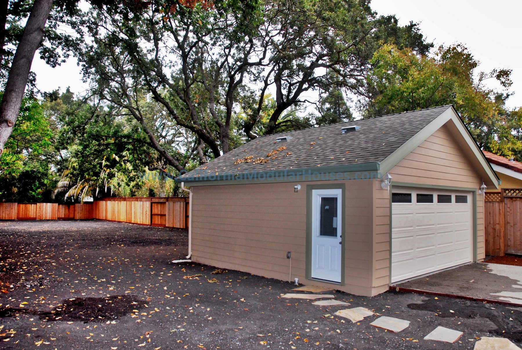 A small beige garage with a white door and a roll-up garage door, set in a backyard with a fenced perimeter and a wooded area with tall trees in the background.