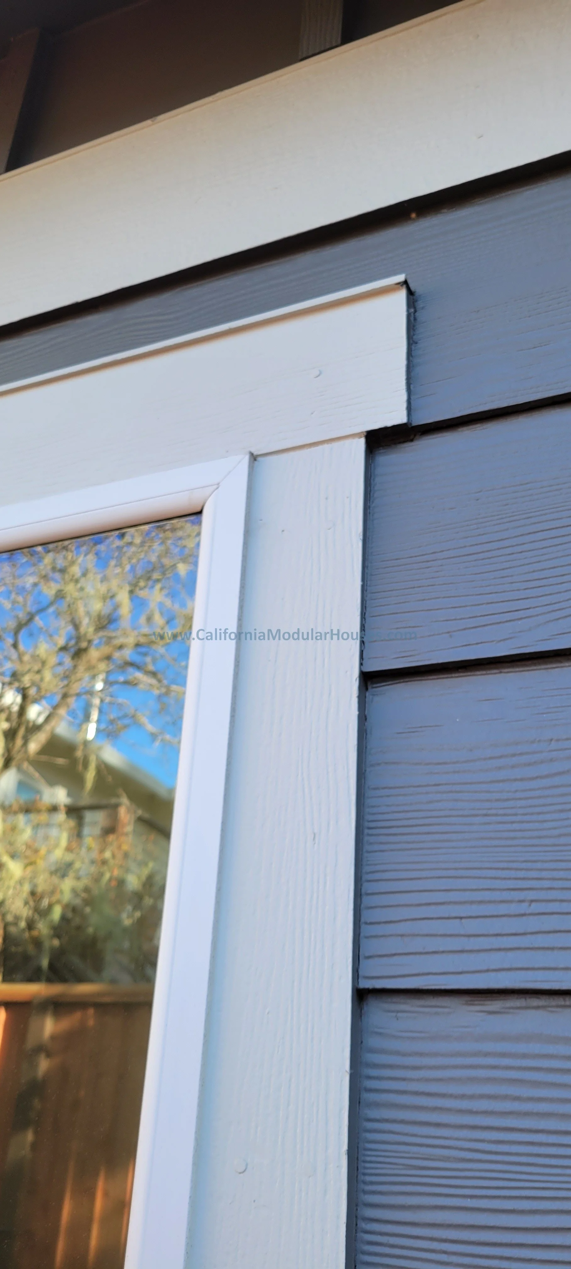 Close-up of a house exterior showing white window trim, gray siding, and a reflection of a tree and blue sky in the window glass.