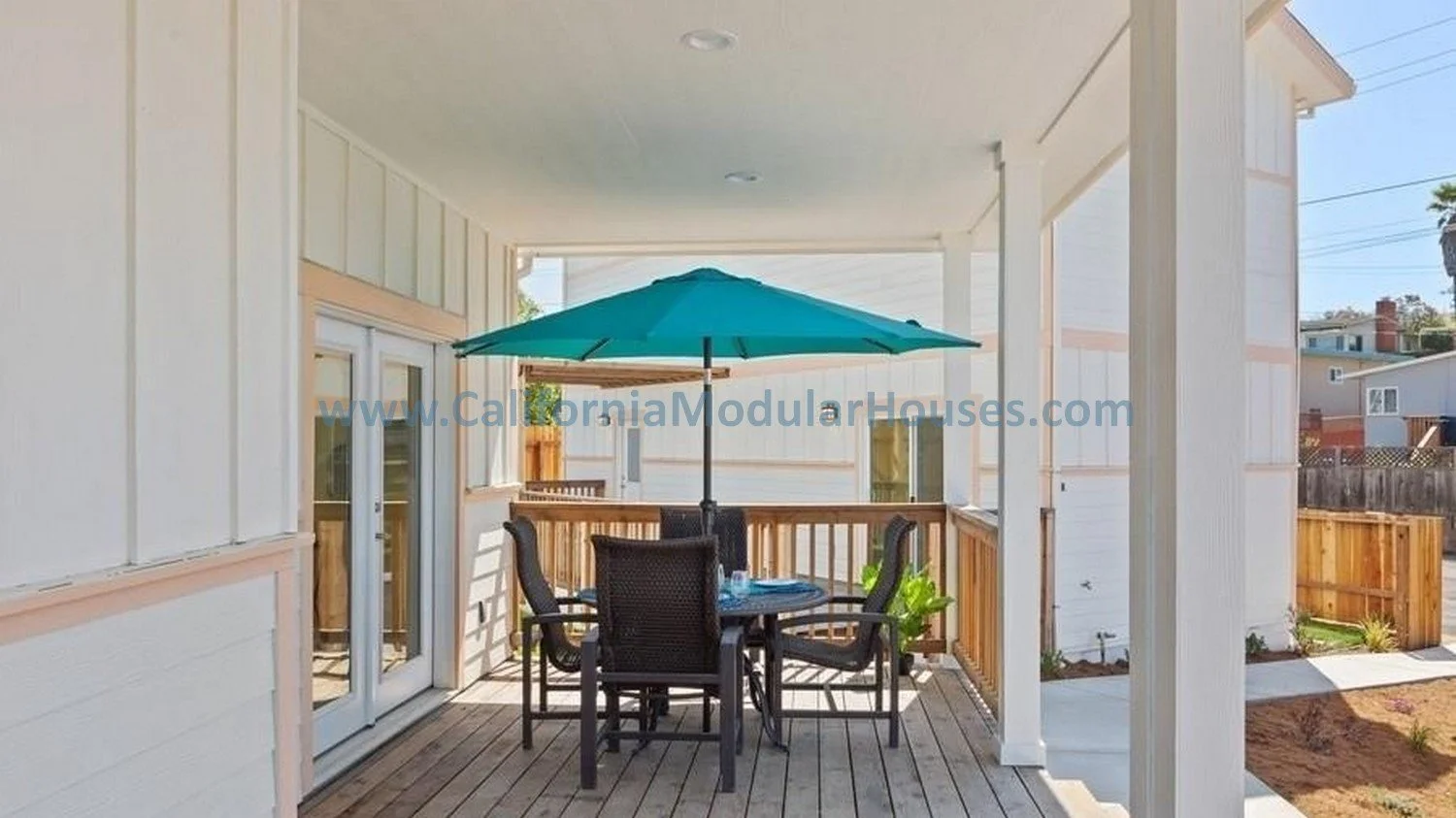 Covered outdoor patio with a round table, four chairs, and a large teal patio umbrella. White house walls with wooden trim and a sliding glass door.  San Francisco Bay Area, CA Two story modular single family modular residence.  Modular home.  