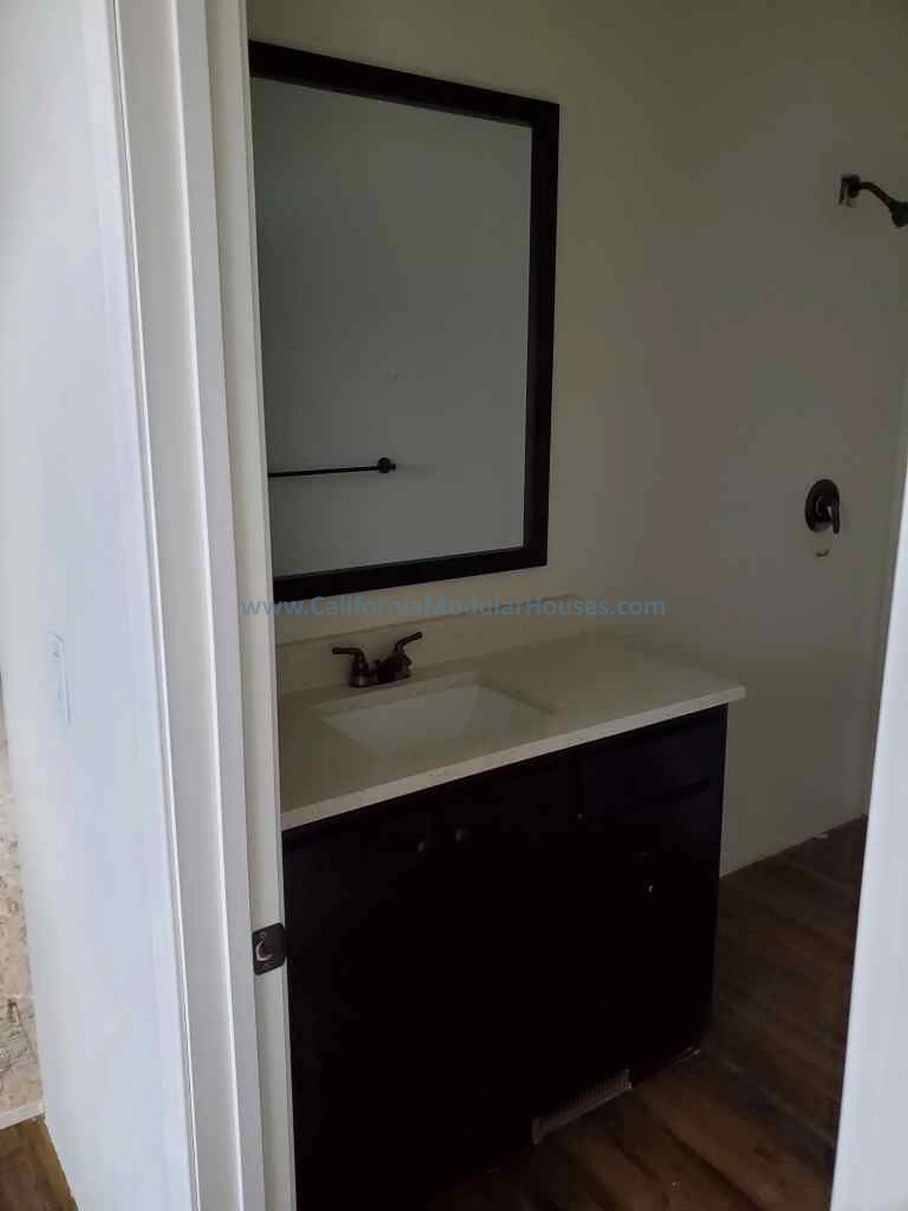 A small bathroom vanity with dark cabinetry and a white countertop, a single sink, and a mirror above it, shown through a doorway.  Two-Story Modular Home. Pre-Fabricated Homes, Prefab Home California,