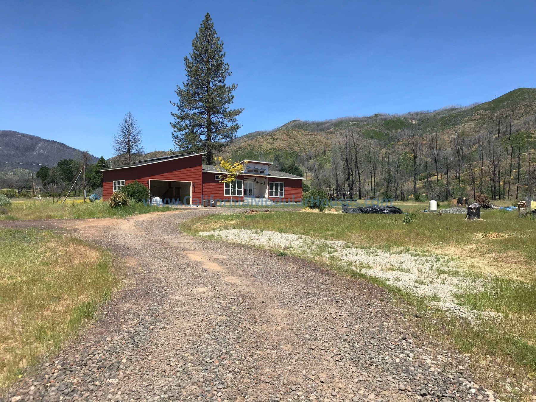 A red barn with white-framed windows is situated on a gravel driveway in a rural landscape with green grass, trees, and mountains in the background under a clear blue sky.  California Prefab Modular Home, California Prefab Modular Home.