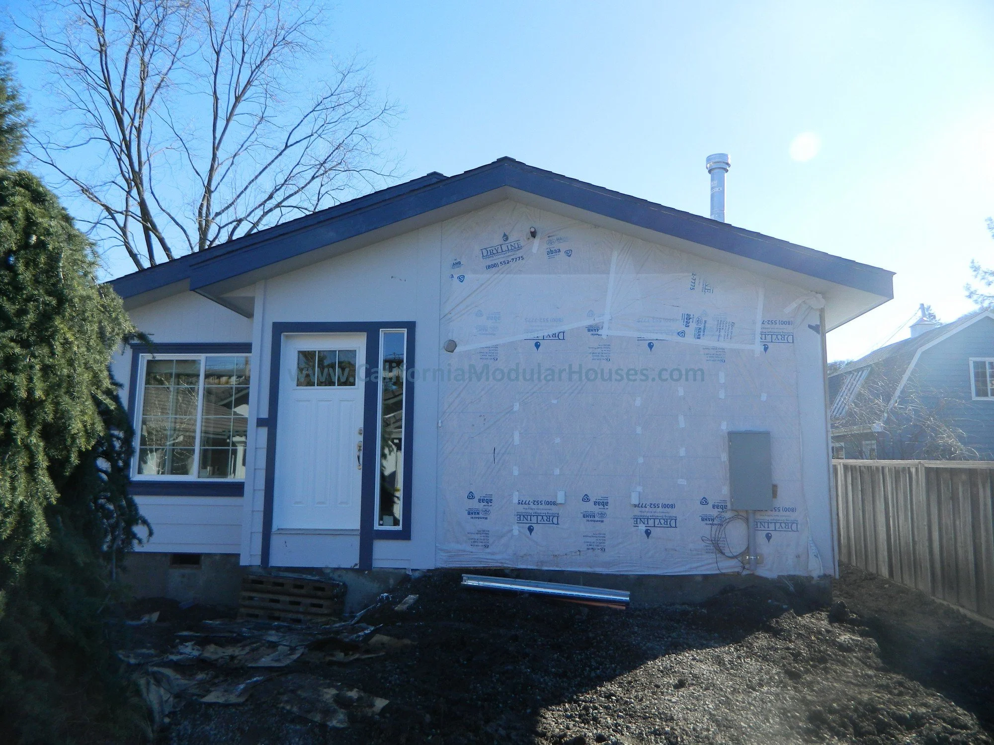 A house under construction with partially finished white siding and an uncovered section revealing insulation. There is a white door with a small window and a large window beside it. The house has a dark blue roof and is surrounded by bare trees and 