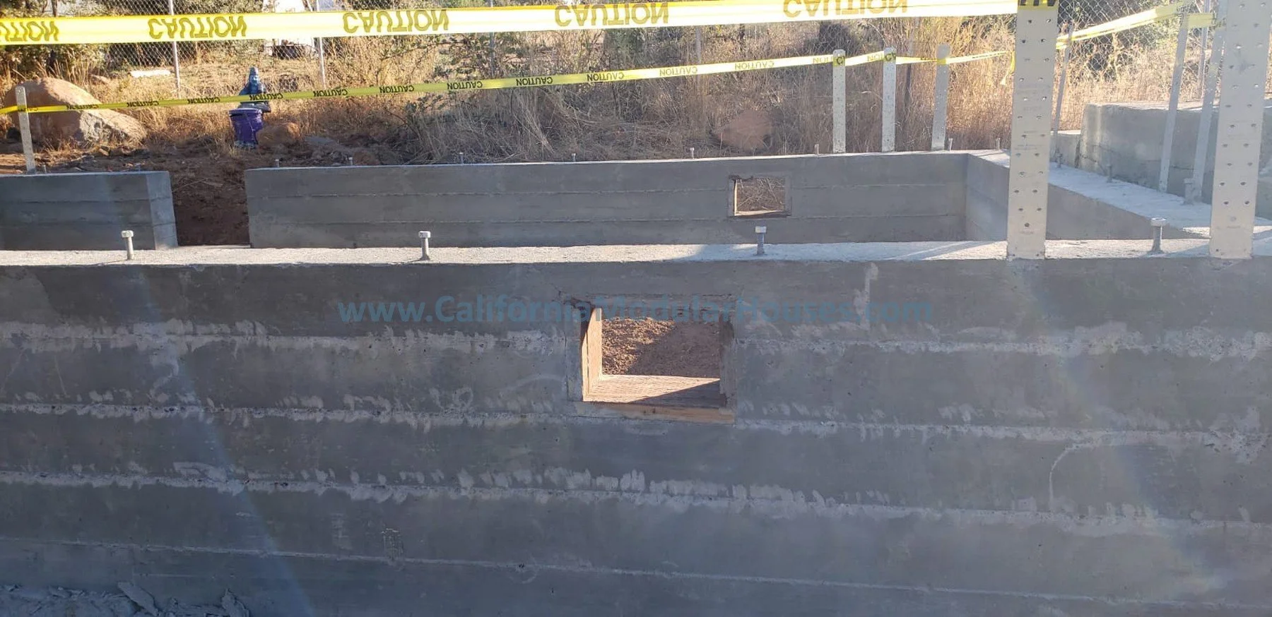 Concrete foundation walls and framing for a building under construction, with yellow caution tape at the top and a small opening in the wall.  Modular Homes, Modular Homes California, Modular Homes in California, Prefab Homes,