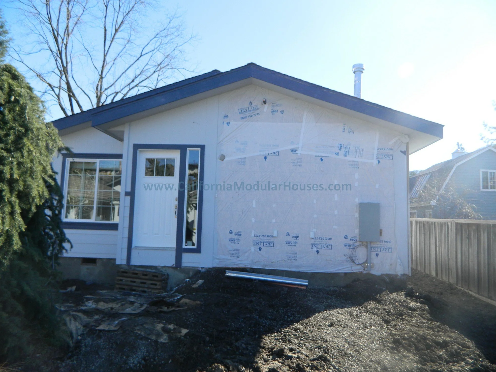 Under-construction house exterior showing partially finished white siding, window, door, and electrical box with wiring, against a clear sky with bare trees.