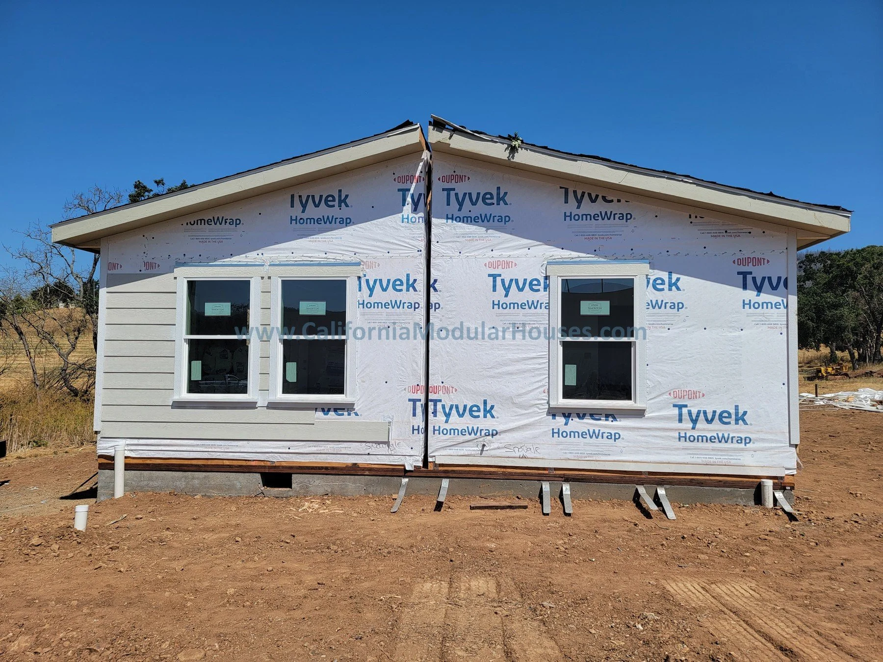 Two-story house under construction with white vinyl siding and two windows on the front, surrounded by dirt and construction materials, under a clear blue sky.