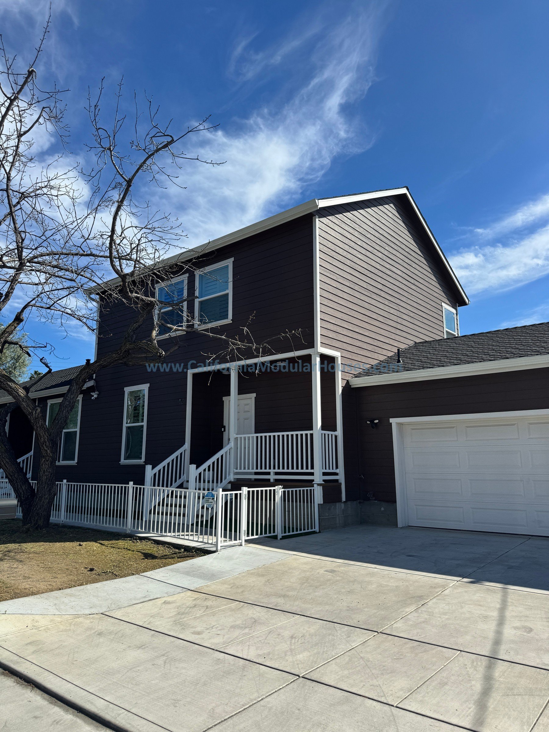 A two-story house with dark brown exterior siding, white trim, and a white porch railing. There is a leafless tree in the front yard, and a concrete driveway leading to an attached garage. The sky is blue with some wispy clouds.