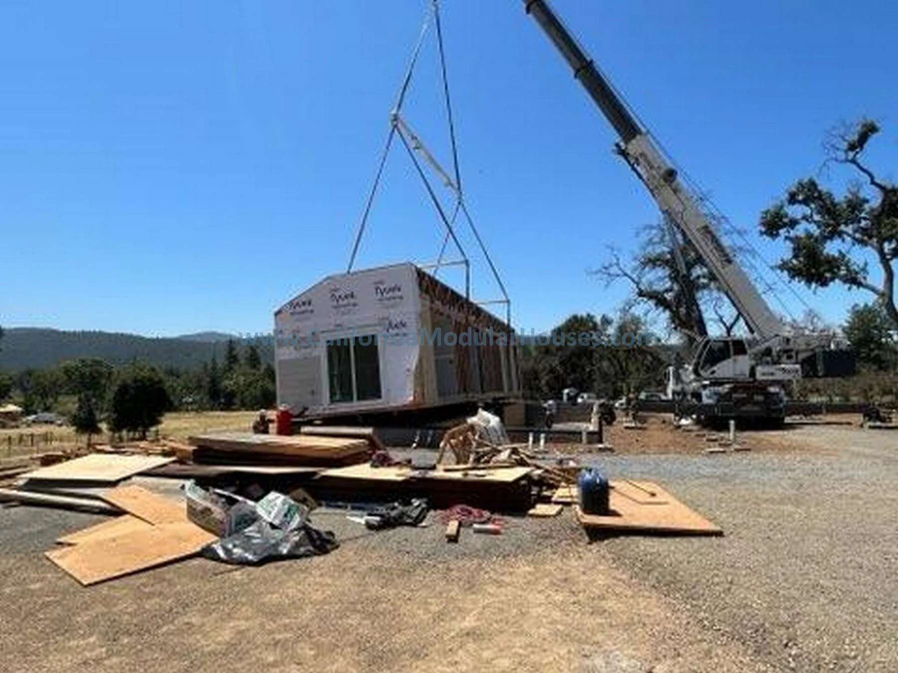 A crane lifting a small prefabricated house onto a foundation at a construction site during daytime. Materials and tools are scattered on the ground, with trees and hills in the background.