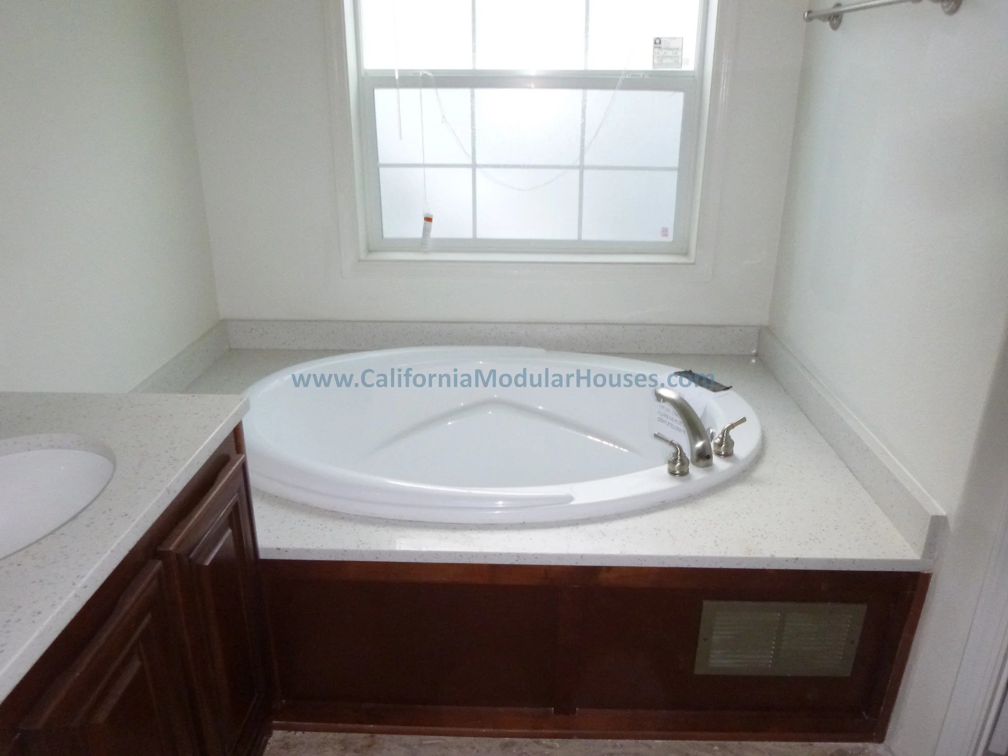 A bathroom with a large corner whirlpool bathtub, a window above it, a marble countertop with a sink on the left, and a wooden cabinet below the bathtub.
