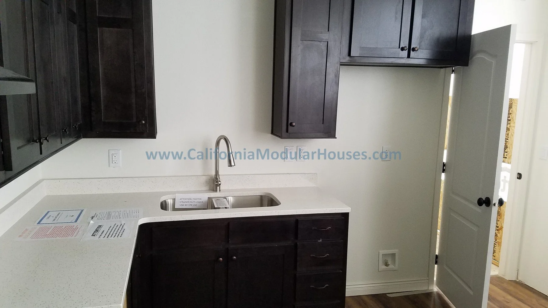 Kitchen with dark wood cabinets, a white countertop, and a stainless steel sink with a faucet. An open door reveals an unfinished room with exposed wall framing.  Modular Homes, Modular Homes California, Modular Homes in California,