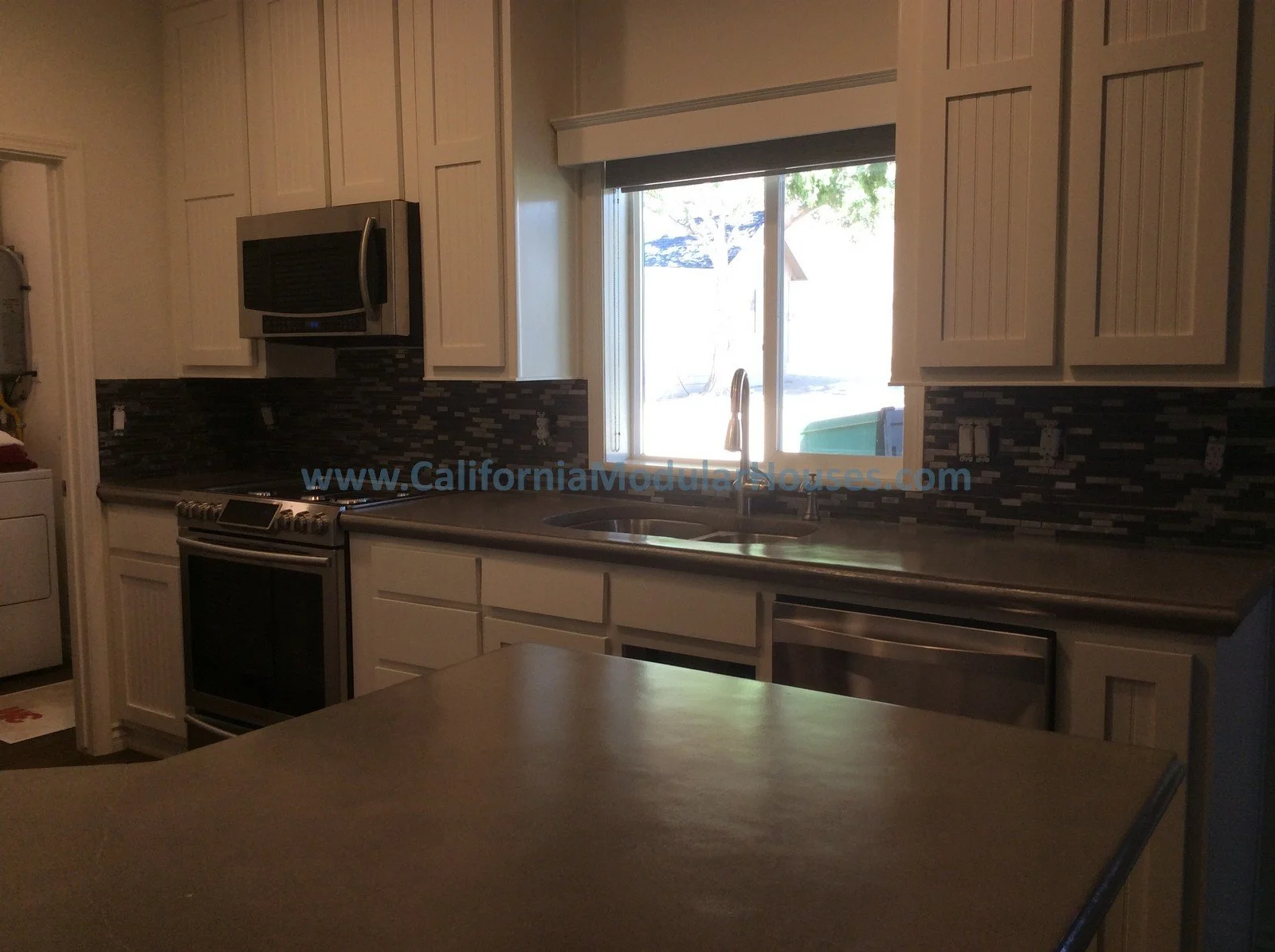 Kitchen with white cabinets, stainless steel stove, microwave, and dishwasher, window with view of trees and backyard, dark mosaic tile backsplash, and light-colored countertops.