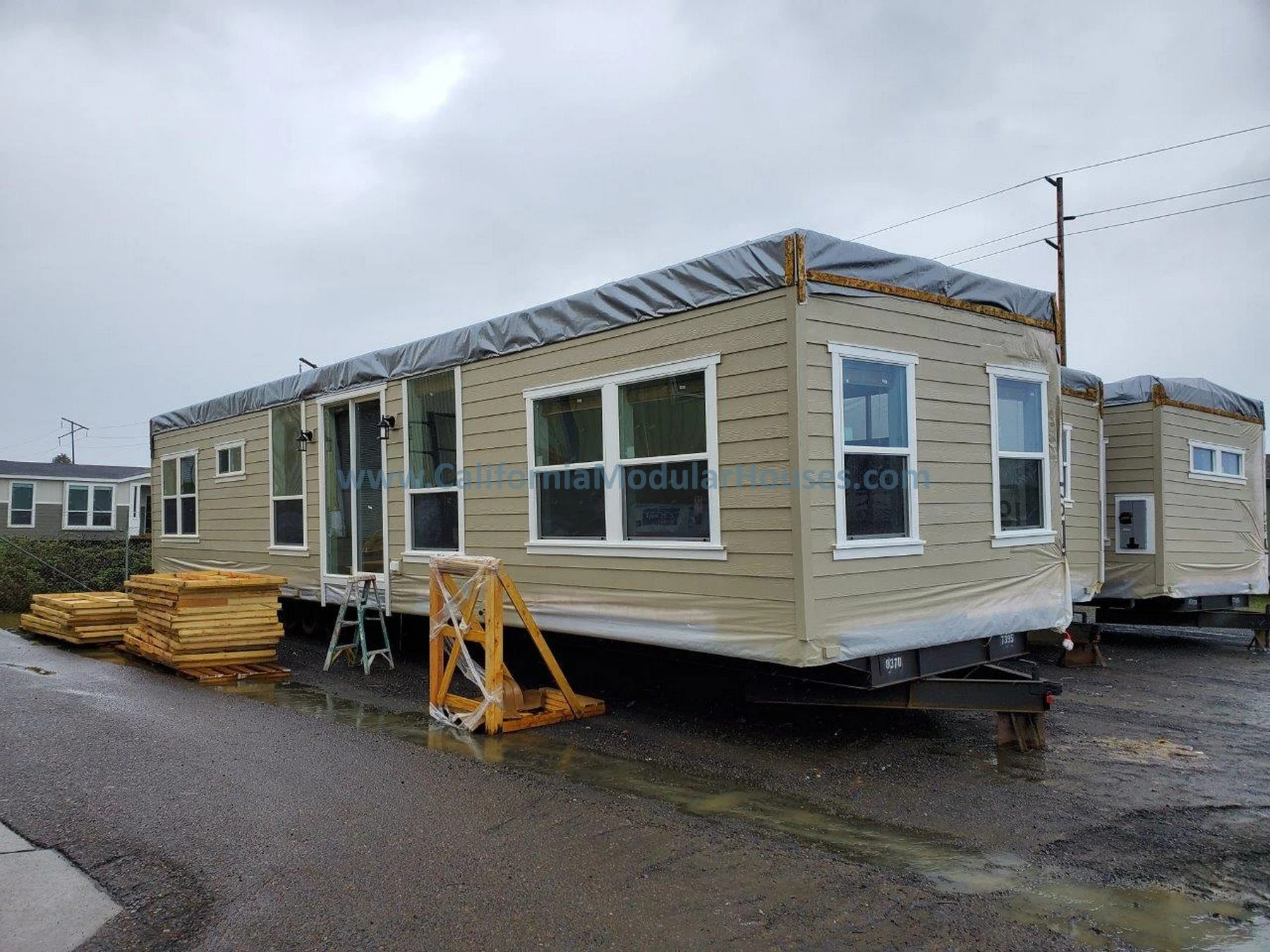 Modularr home under construction with beige siding, multiple windows, and a gray tarp covering its roof with stacked lumber nearby.  Modular Homes in California, Prefab Home California, Pre-Fabricated Homes, Two-Story Modular Home.