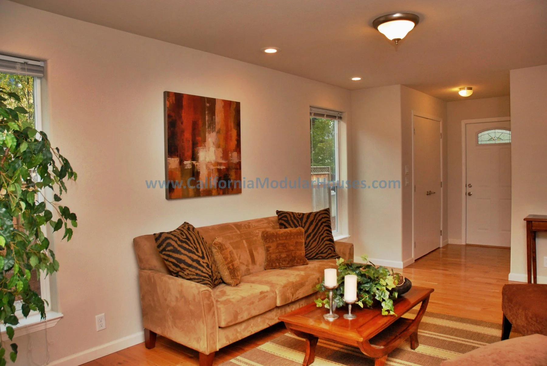 Living room with a beige sofa, dark animal print pillows, a wooden coffee table with candles and greenery, a window with blinds, a colorful abstract wall art, hardwood floors, and a ceiling light fixture.
