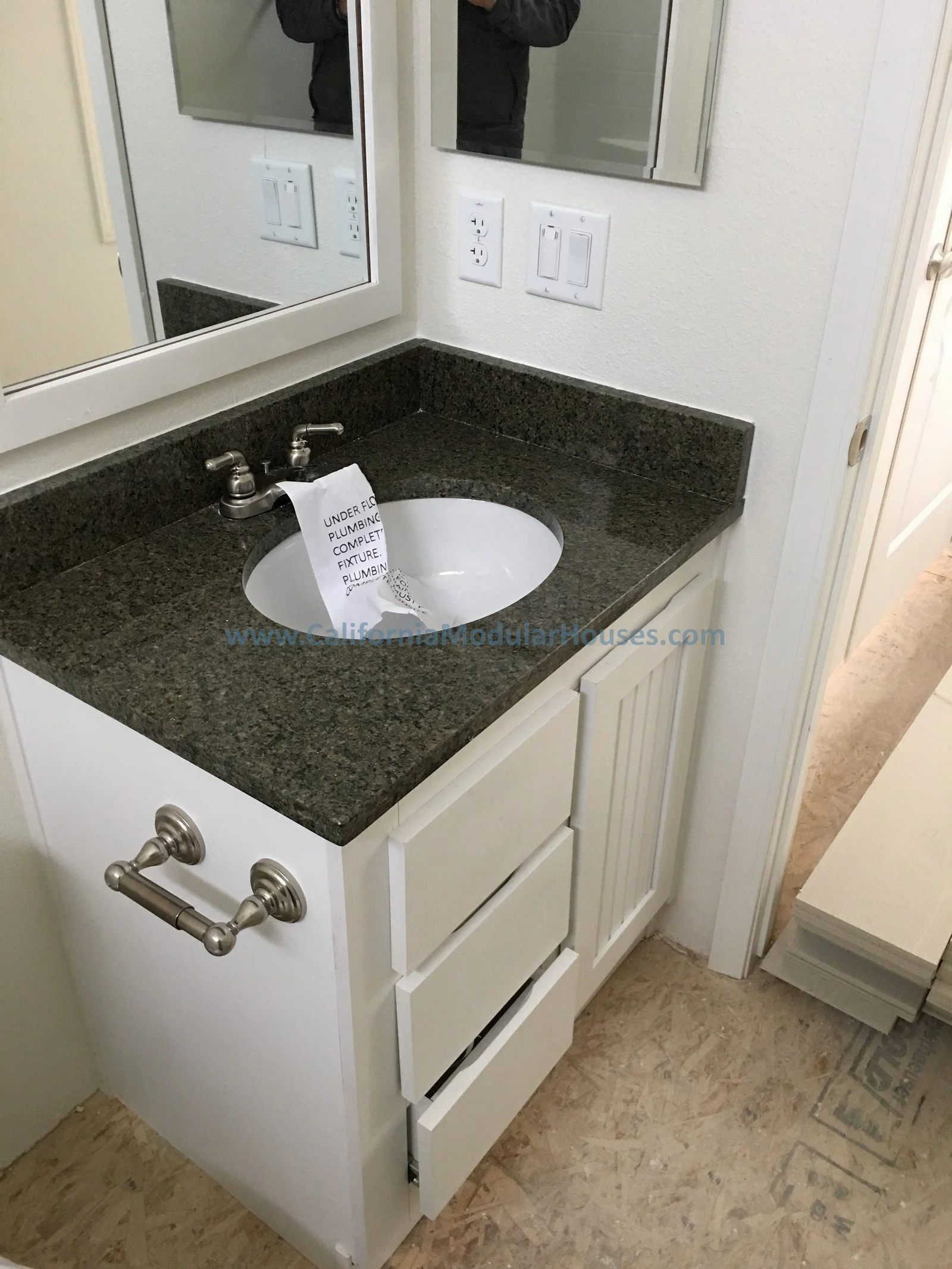 Bathroom vanity with a dark granite countertop, white cabinet with drawers and closet door, and a mirror above the sink. A sign hanging in the sink indicates plumbing is incomplete.