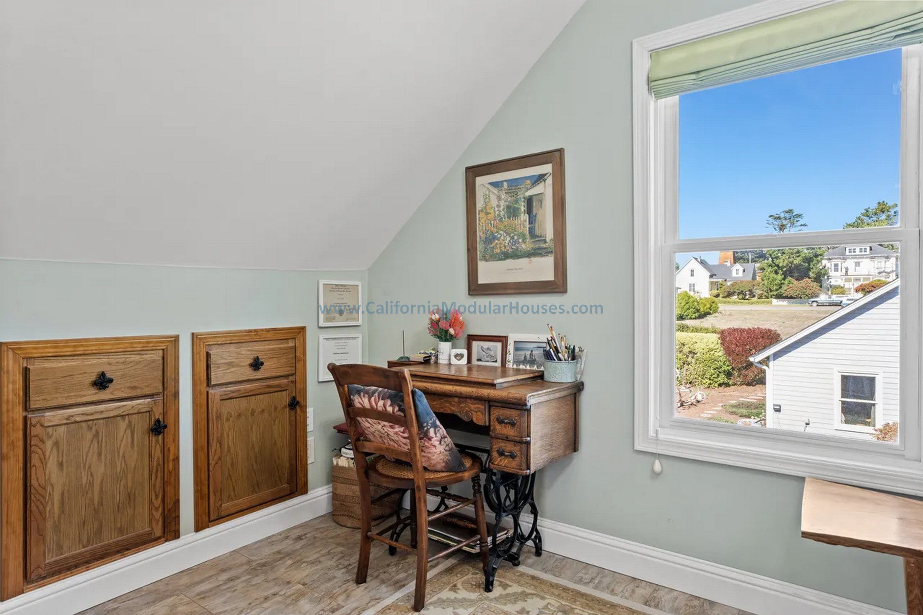 Small home office with a vintage wooden desk and chair, a window with a view of neighboring houses and greenery, and light green walls decorated with artwork and framed certificates.