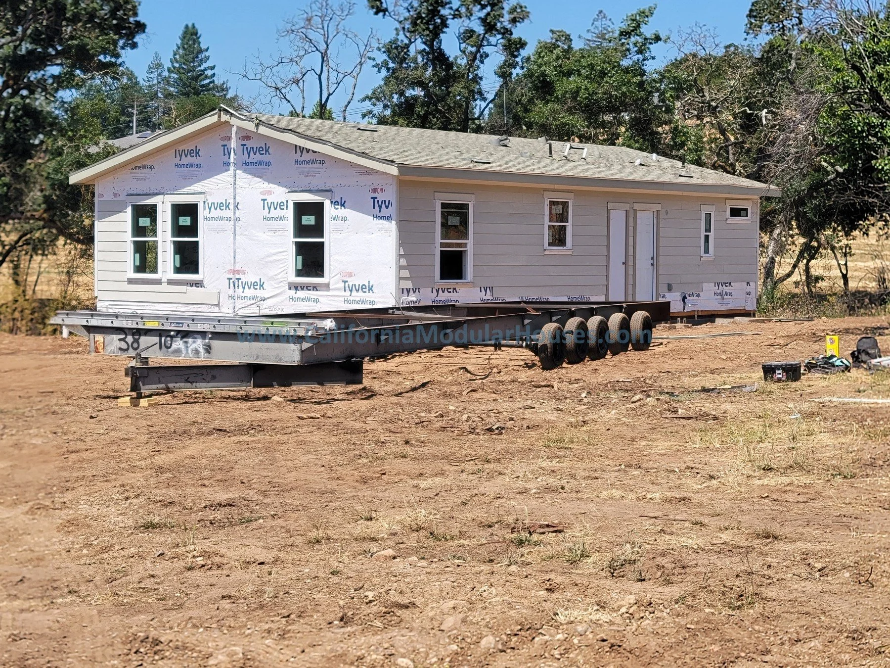 A house under construction on a trailer in an open, dirt area with trees in the background.