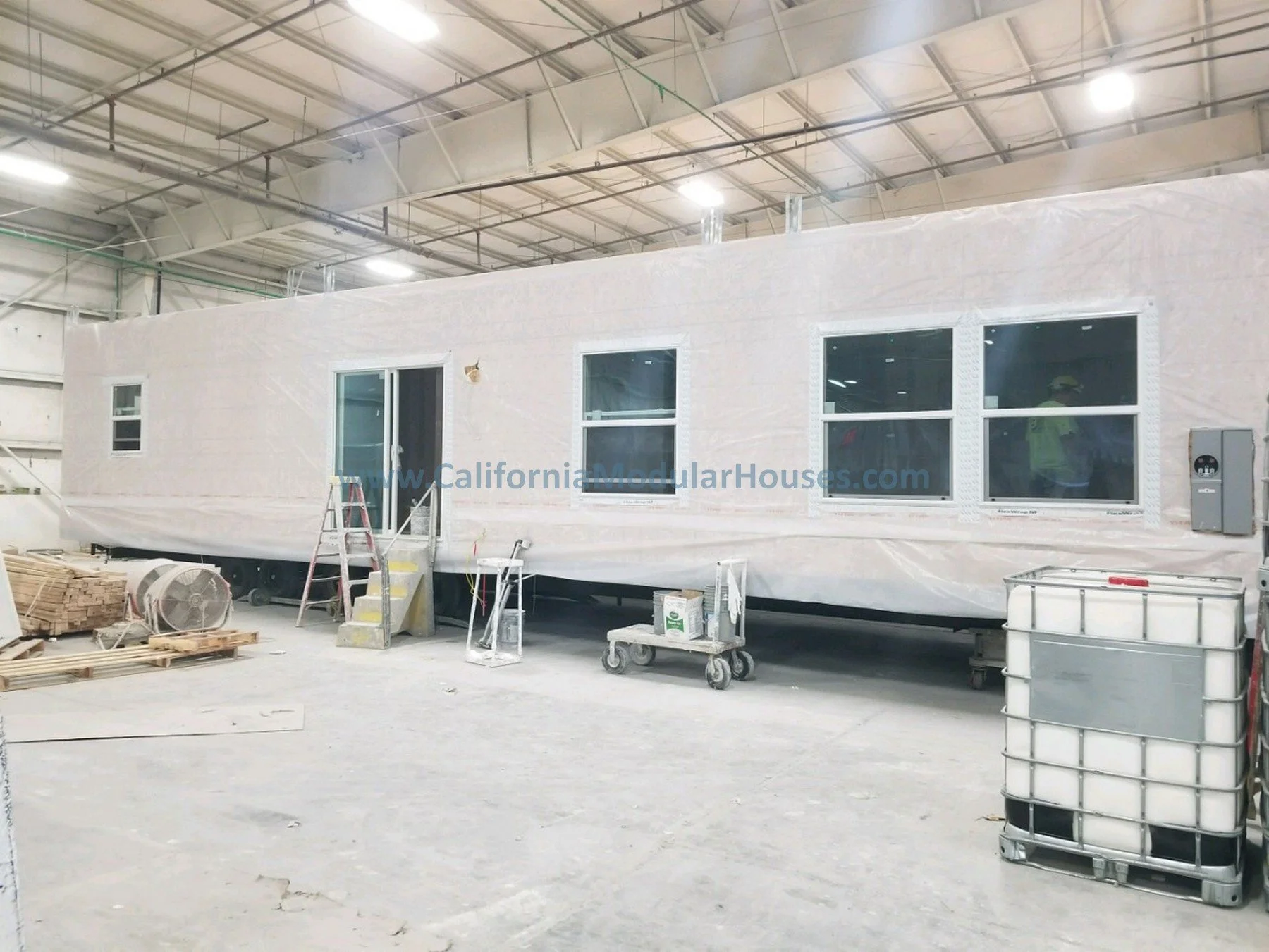 Incomplete mobile modular house under construction inside the factory, with windows and door installed, surrounded by construction equipment and materials.  Two-Story Modular Home. Pre-Fabricated Homes,