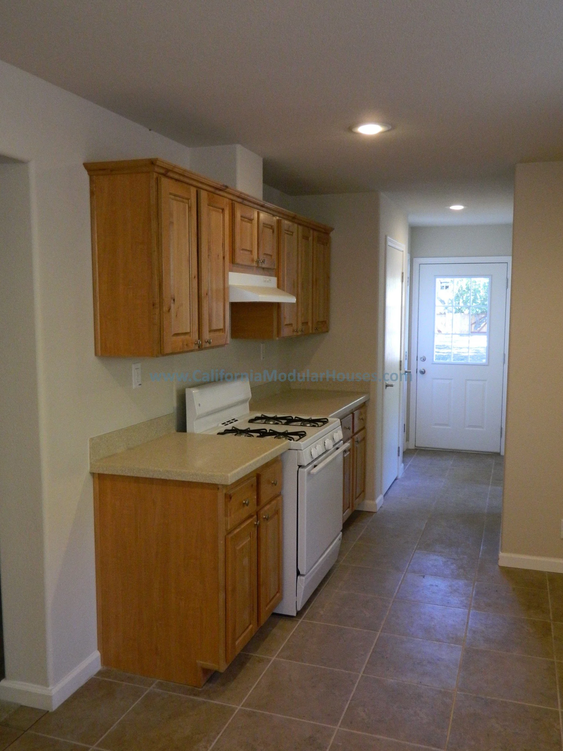 A small kitchen with wooden cabinets, a white stove, and a countertop with a beige surface, leading to a door outside.