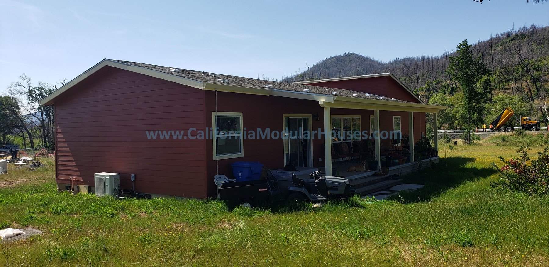 Red house with a porch, surrounded by green grass and trees, with a mountain in the background. A cement mixer truck is visible in the distance.  California Modular Houses,  Middletown Modular Home.