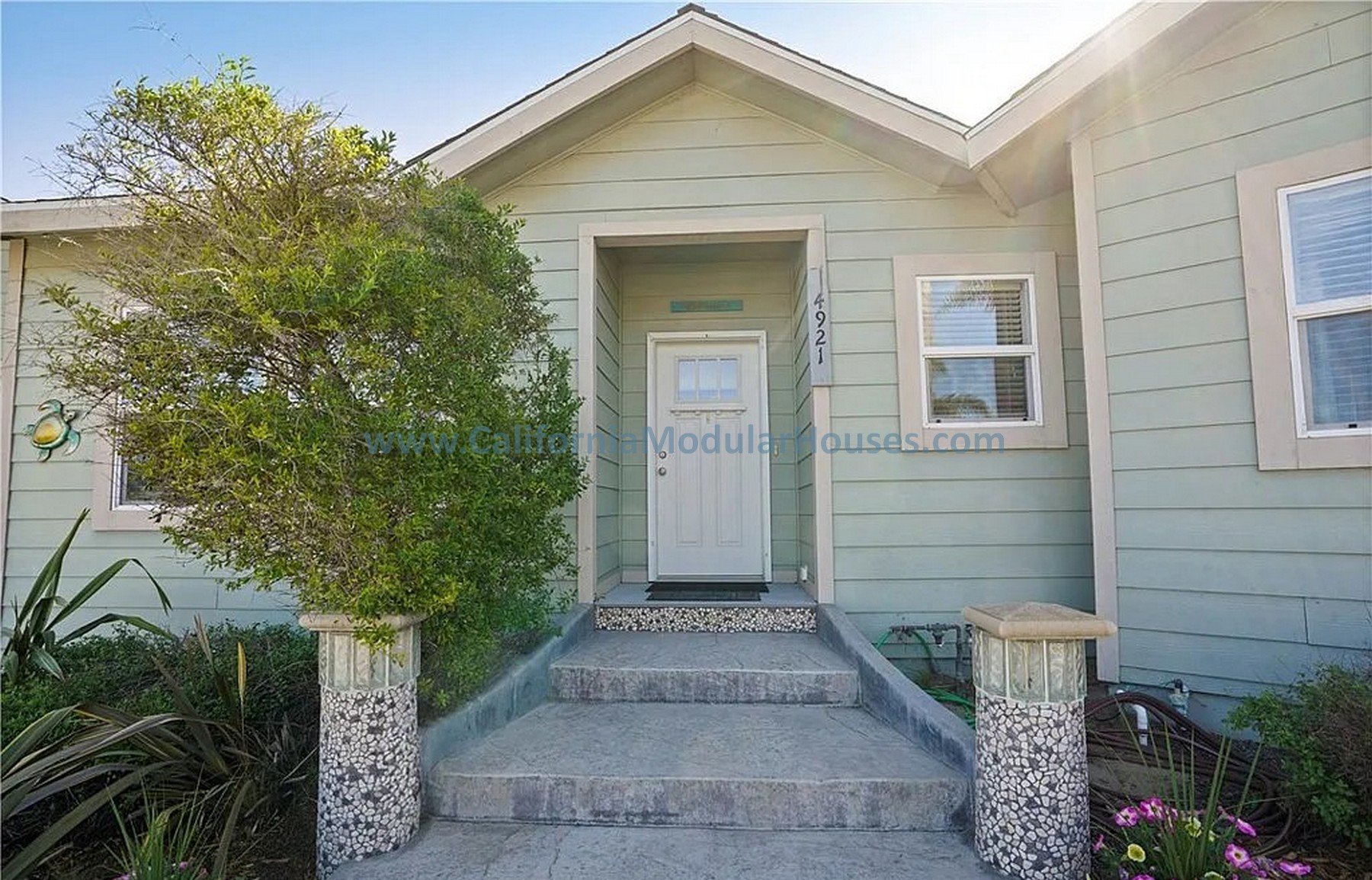 Front entrance of a light green house with white trim, featuring a white front door, two windows, a small porch with concrete steps, and decorative mosaic columns on either side of the steps.   California Modular Houses. Modular home construction Cal