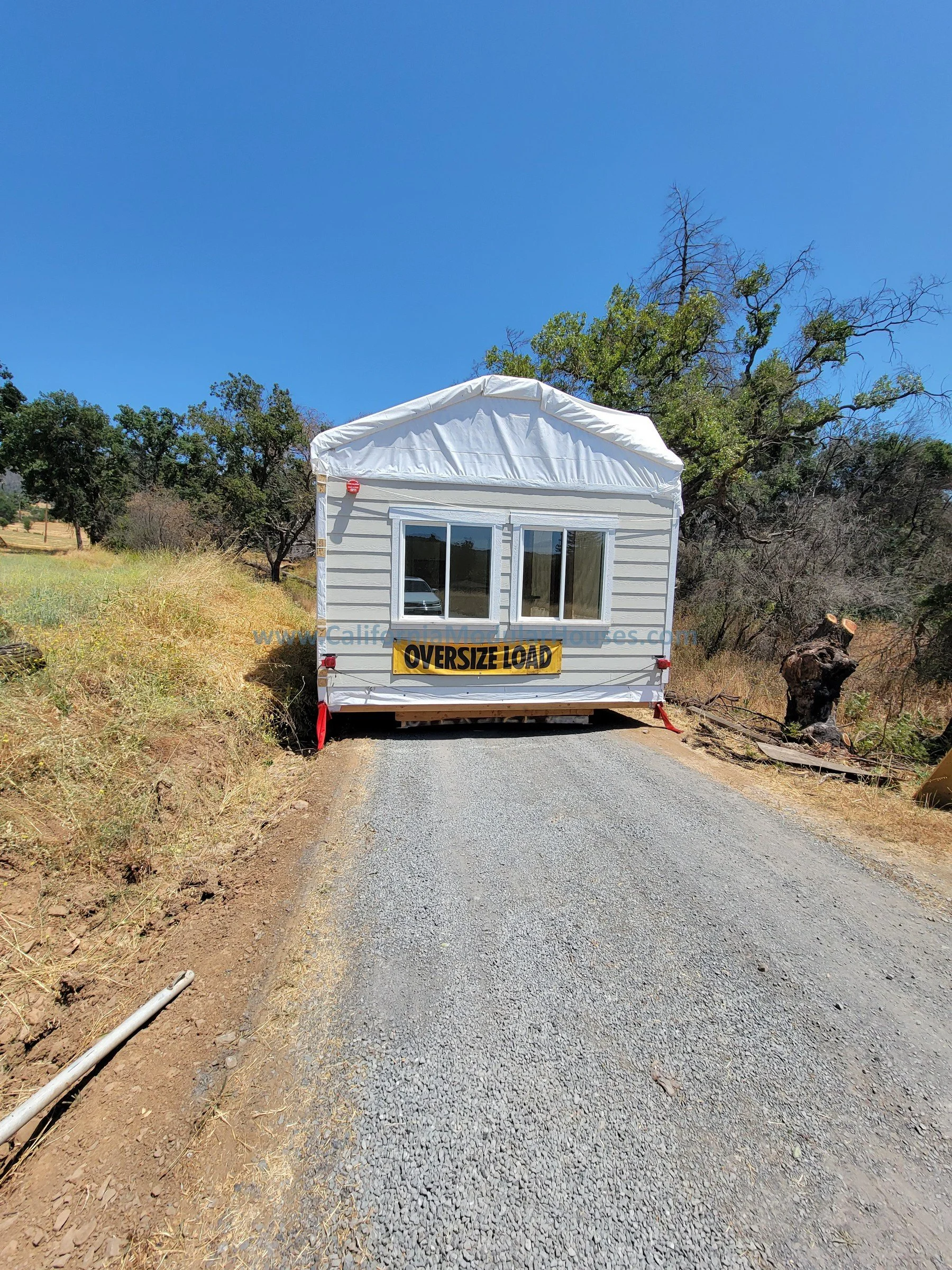 A small white mobile home with a sign that reads 'OVERSIZE LOAD' is blocking a rural gravel road, surrounded by dry grass, trees, and clear blue sky.
