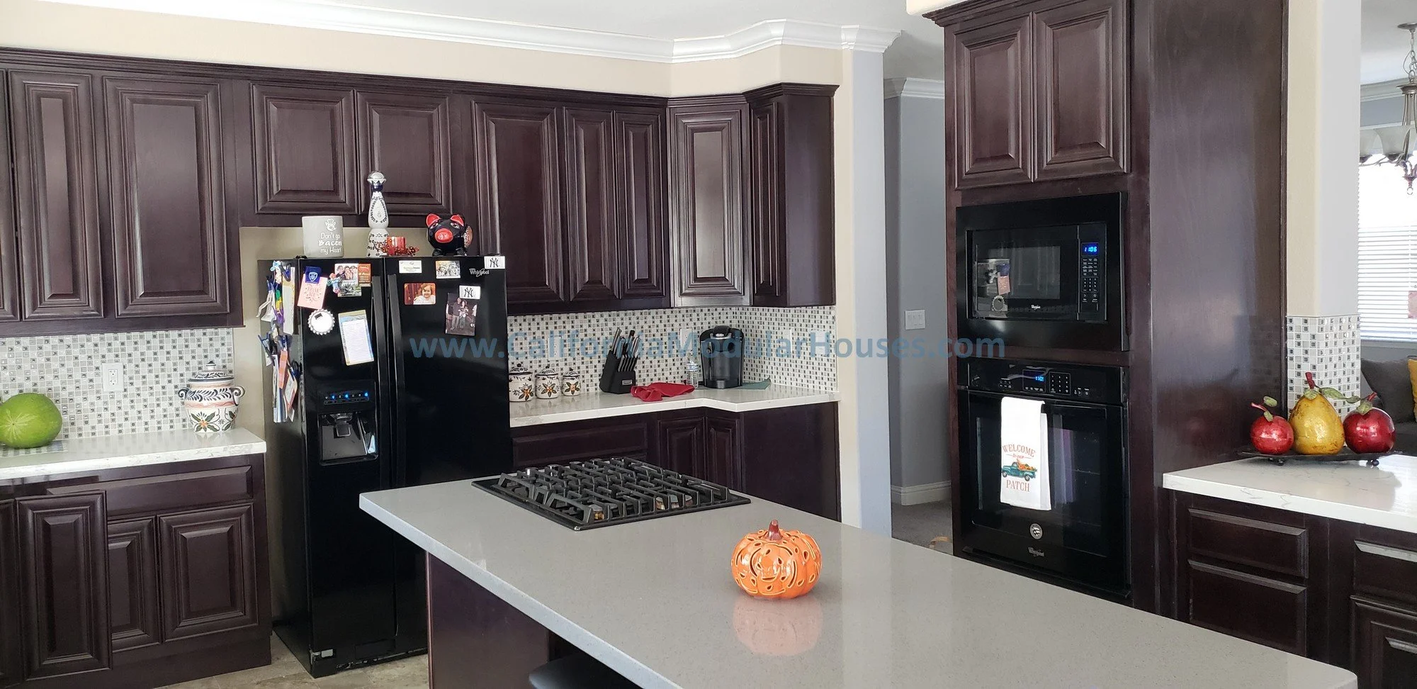The kitchen has dark wood cabinets, a white countertop island, a black refrigerator.  It features a built-in oven and microwave.  The cooktop in the over-sized island is extremely functional in this modular home.   