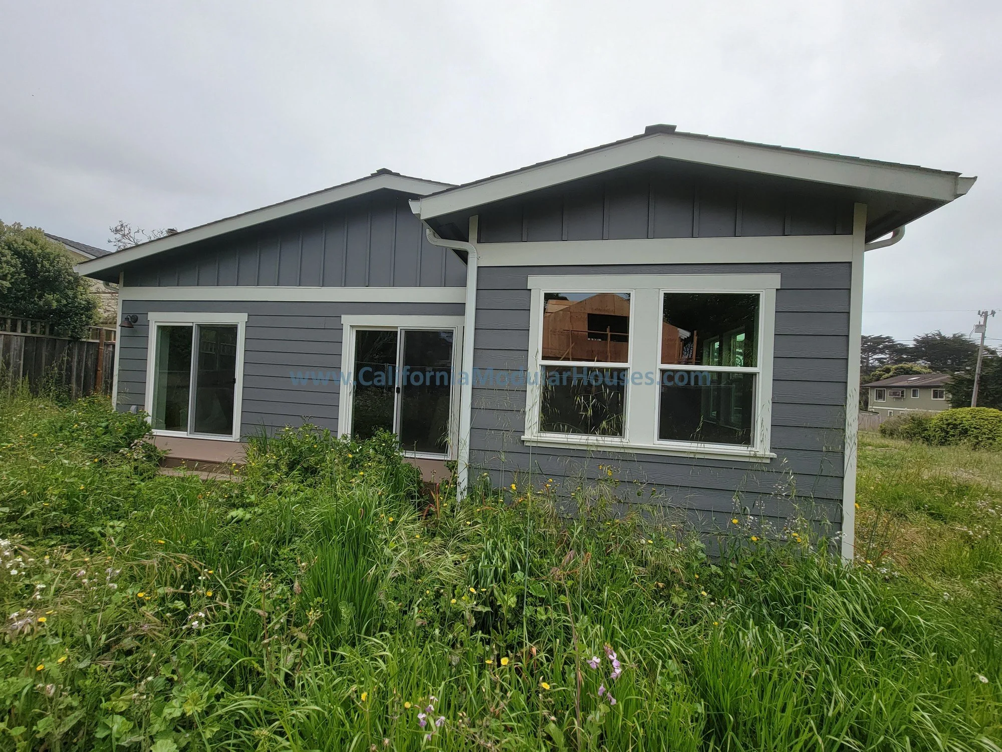 A new gray house with white trim under an overcast sky, surrounded by tall grass and wildflowers.