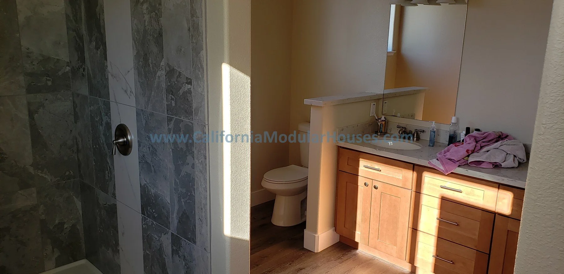Bathroom with a wooden vanity, marble countertop, and a large mirror. To the left, a separate shower area with gray and white tiled walls. The bathroom has a yellow wall and a window letting in sunlight.  Prefab Modular ADU