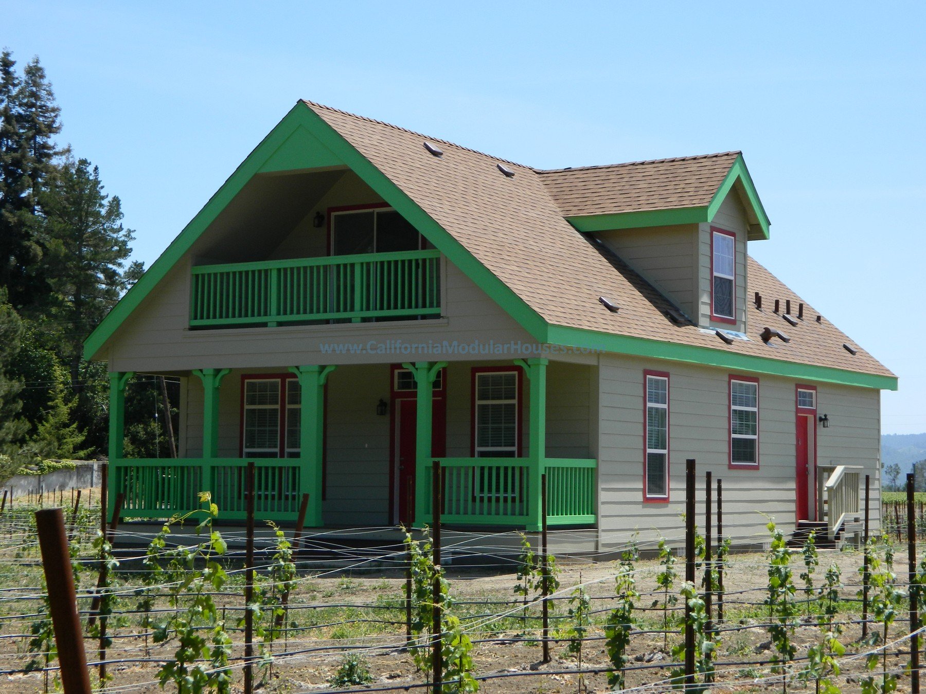 A two-story house with beige siding and green trim, a balcony on the upper floor, and a front porch with green railings, surrounded by a vineyard with rows of grapevines.