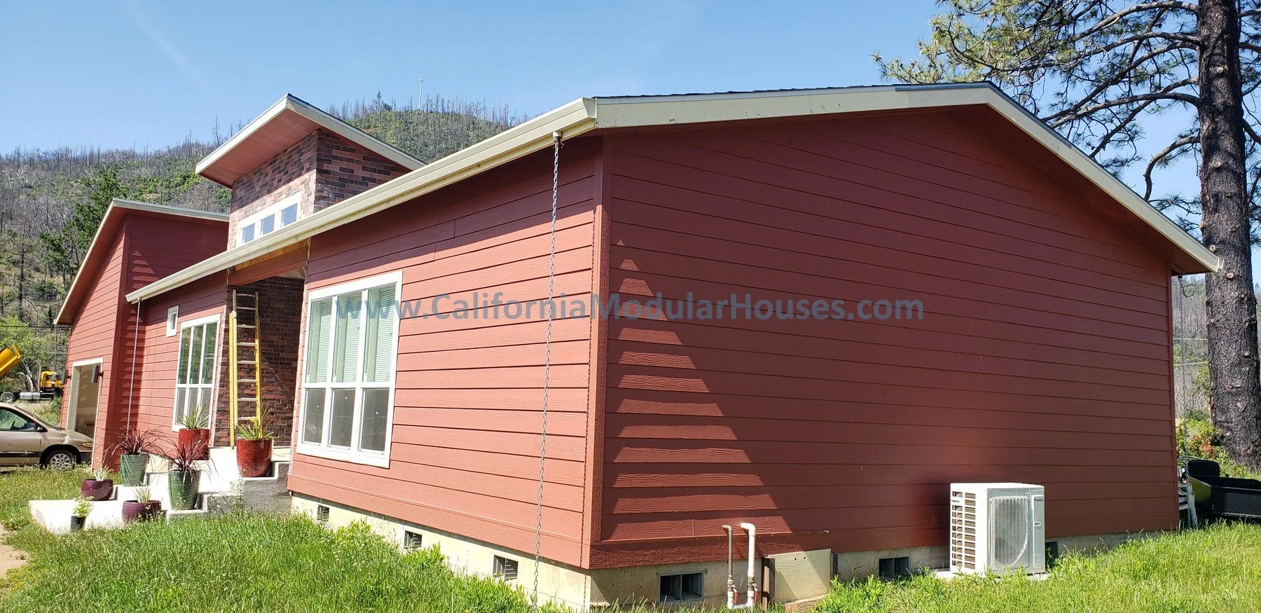 A modern red modular house with white window frames and a small chimney, situated on a grassy yard with potted plants, trees, and a hillside in the background. Prefab Modular Homes California, California Modular.