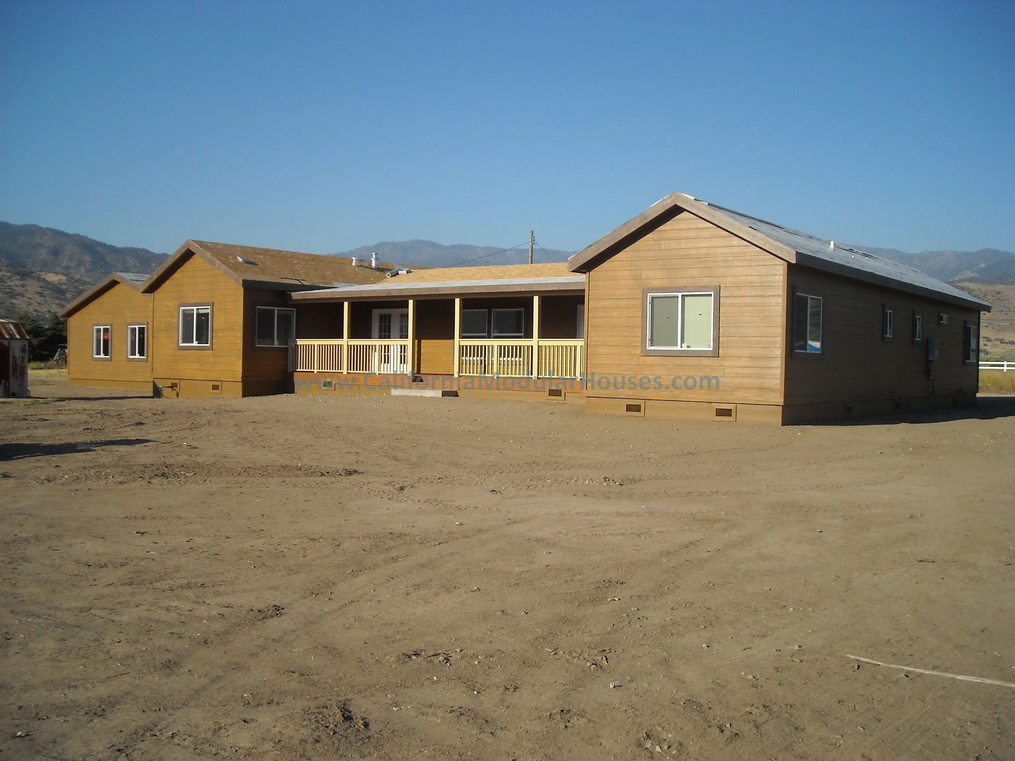 A wooden house with a central porch, multiple windows, and a gabled roof, situated in a dry, dirt area with mountains in the background.