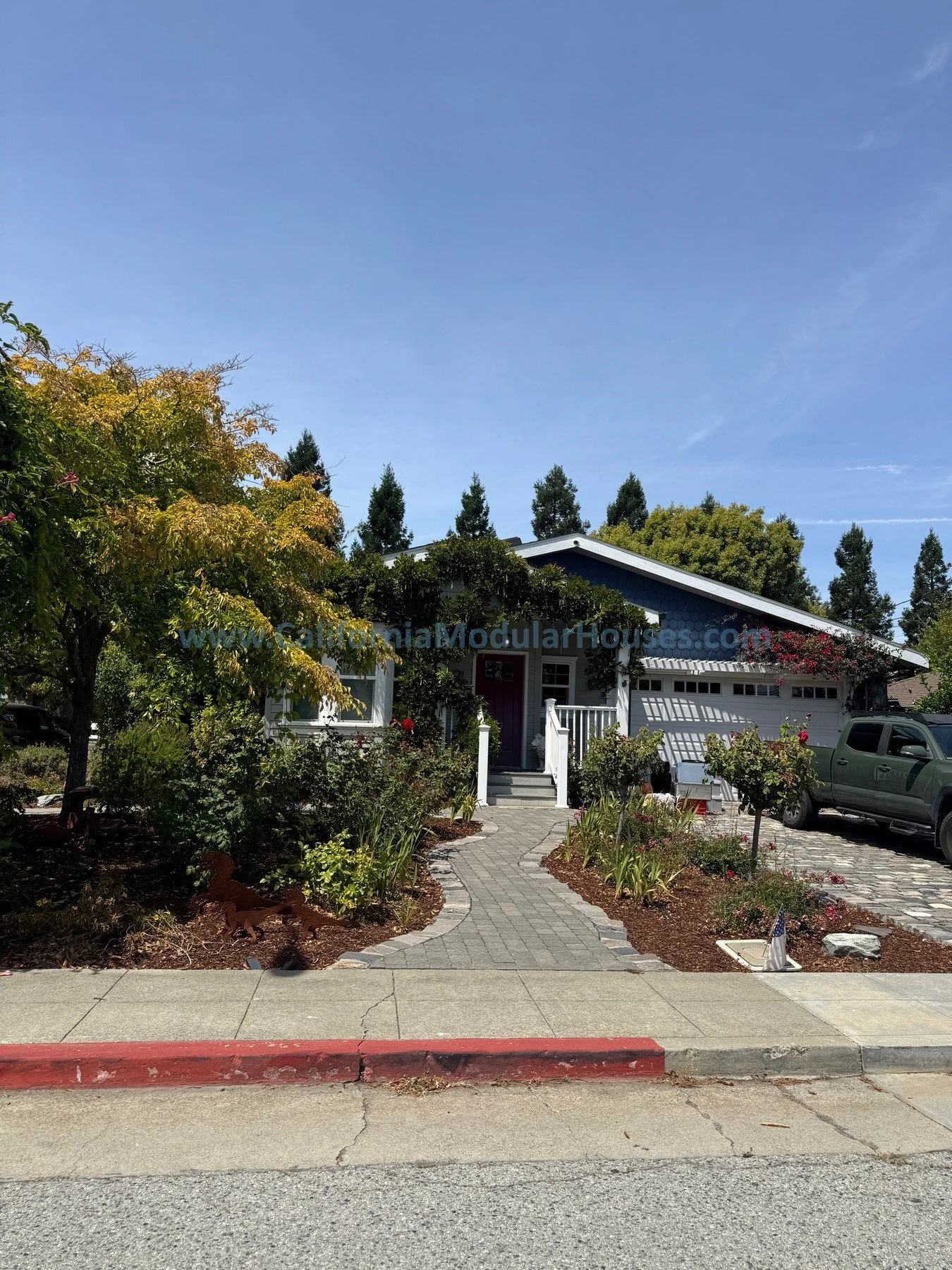 A house with a garden in front, featuring a concrete pathway and lush plants. The house has a dark blue exterior, white trim, and a white garage door. There are multiple trees and shrubs around, with a red door at the entrance and a silver car parked