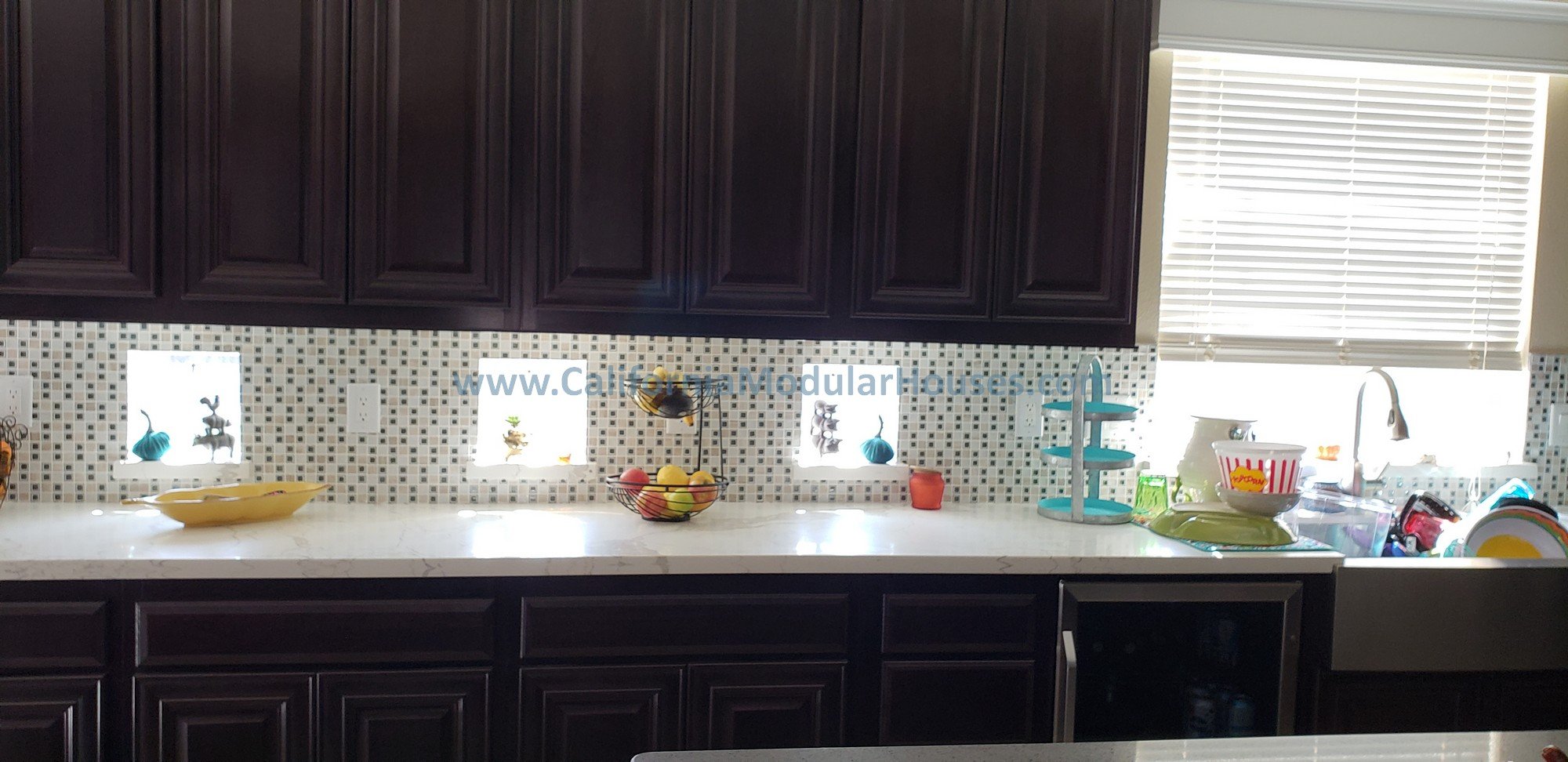 Kitchen counter with dark wood cabinets above, a mosaic tile backsplash, and a window with blinds.  The designer added in these windows under the cabinets to maximize natural lighting on behalf of the client of this modular home.  