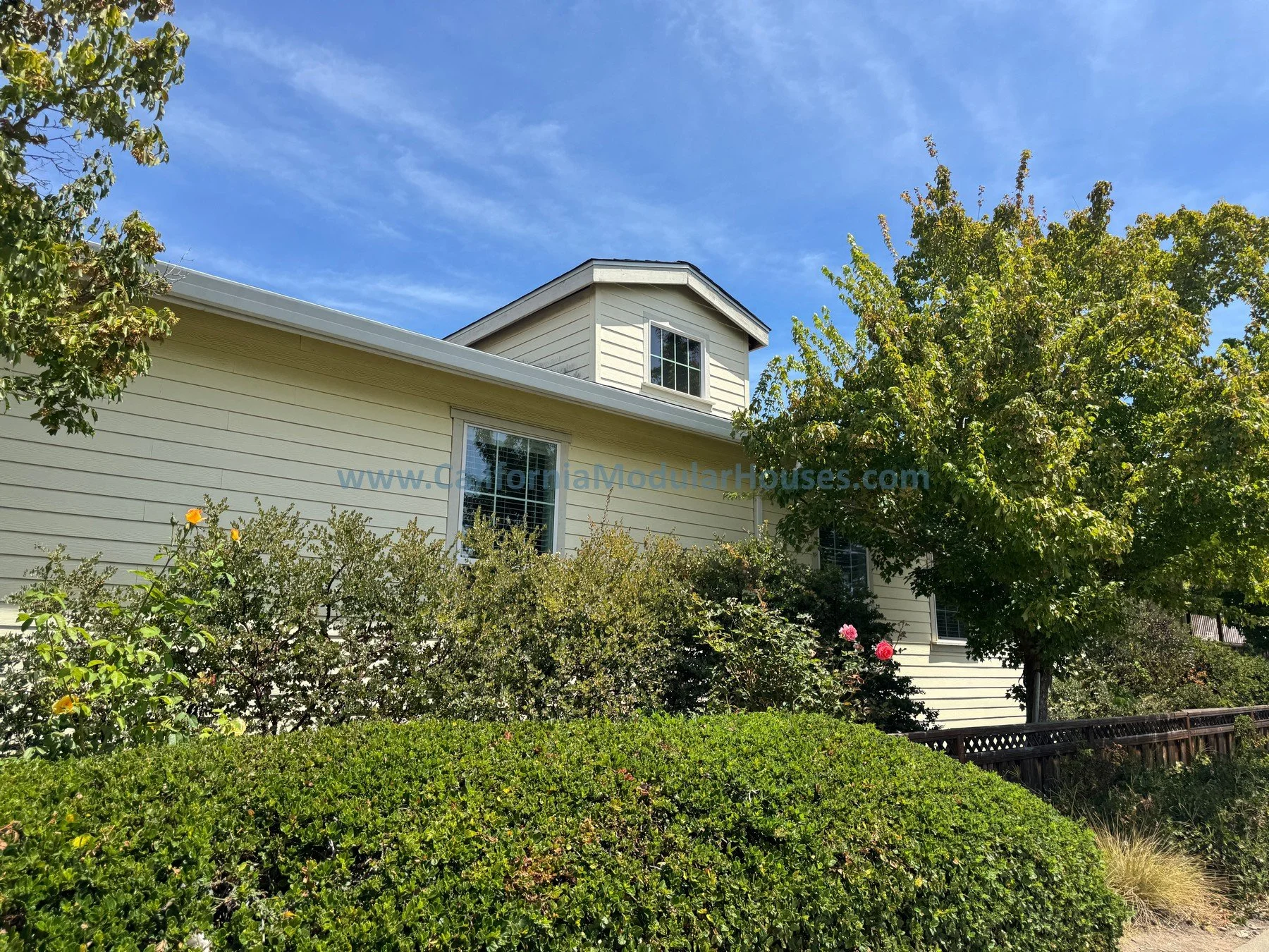 A two-story house with beige siding, surrounded by trees and shrubs, under a clear blue sky.