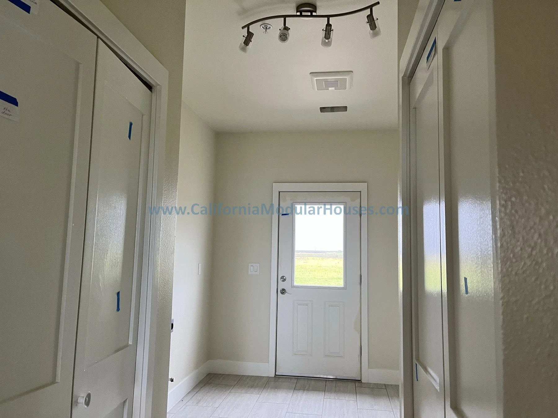 Interior view of a room with white doors, walls, and tiled floor, a window and door at the back, ceiling lights, and electrical outlets.