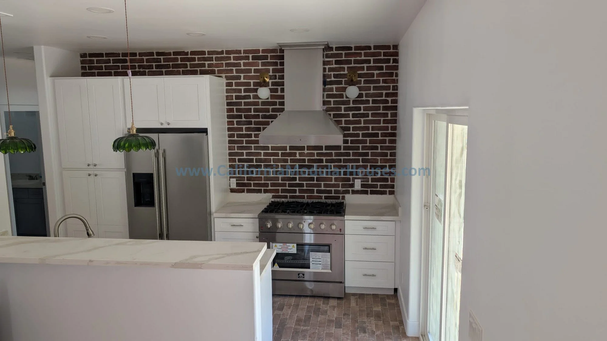 Kitchen with white cabinets, stainless steel refrigerator, brick accent wall, stainless steel stove, white countertops, and two green pendant lights.