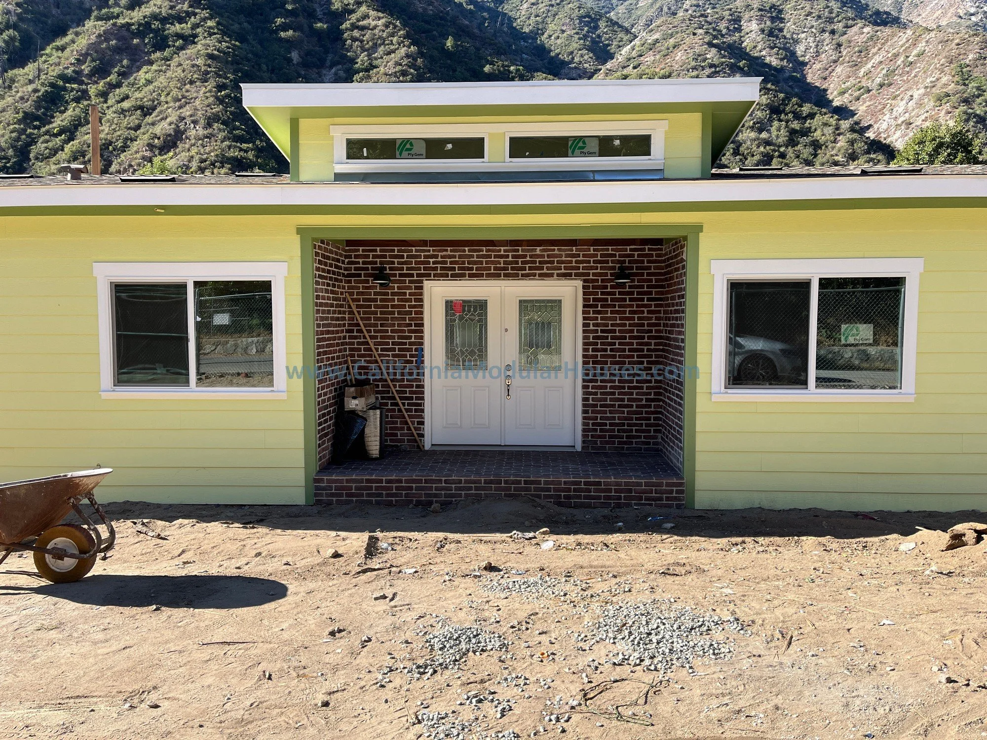 Newly constructed house with yellow and brick exterior, two white front windows, white front door, and unfinished dirt yard.