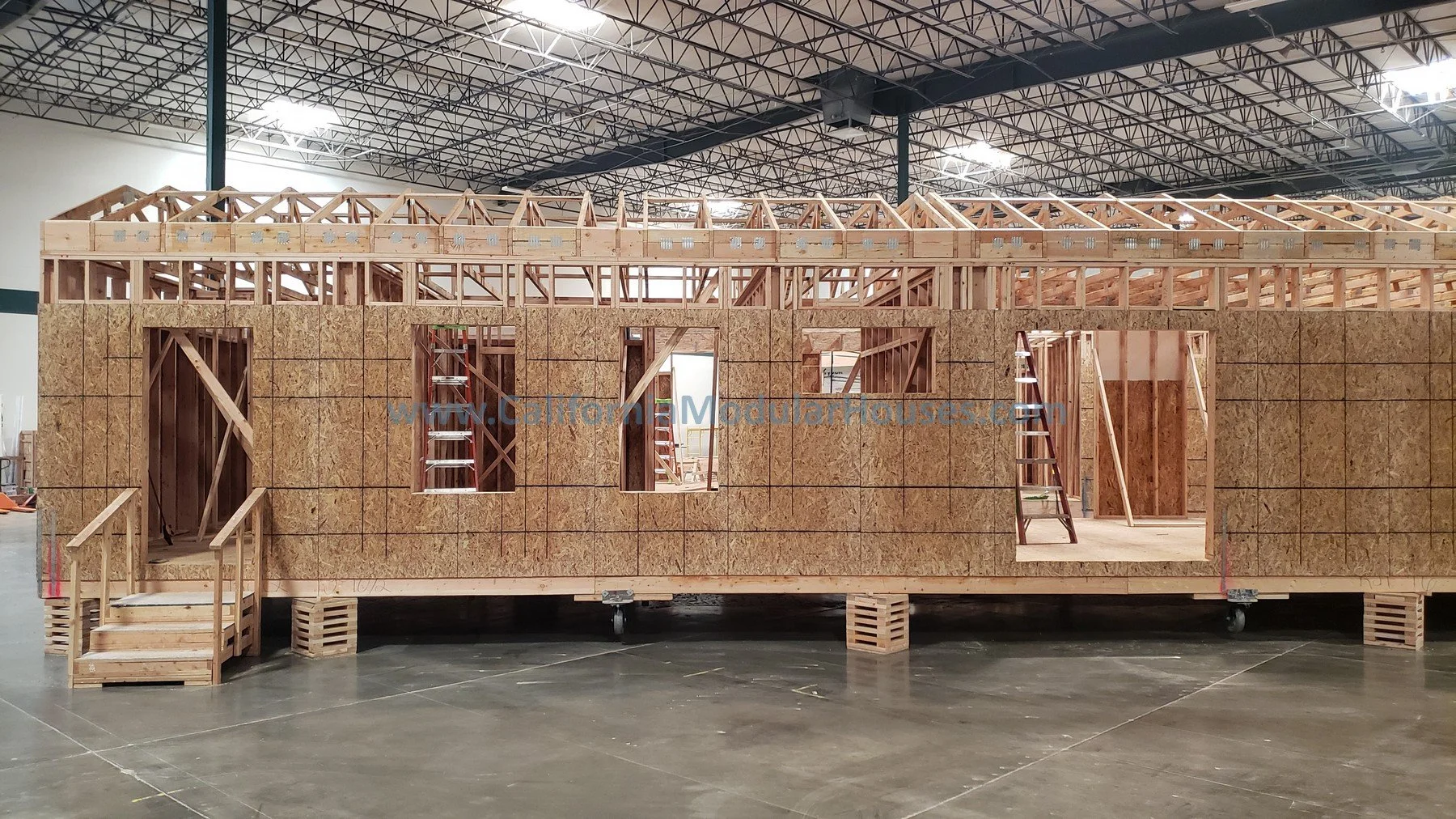 Interior view of a partially built modular house framework with wooden walls, windows, and stairs, inside a large industrial warehouse.