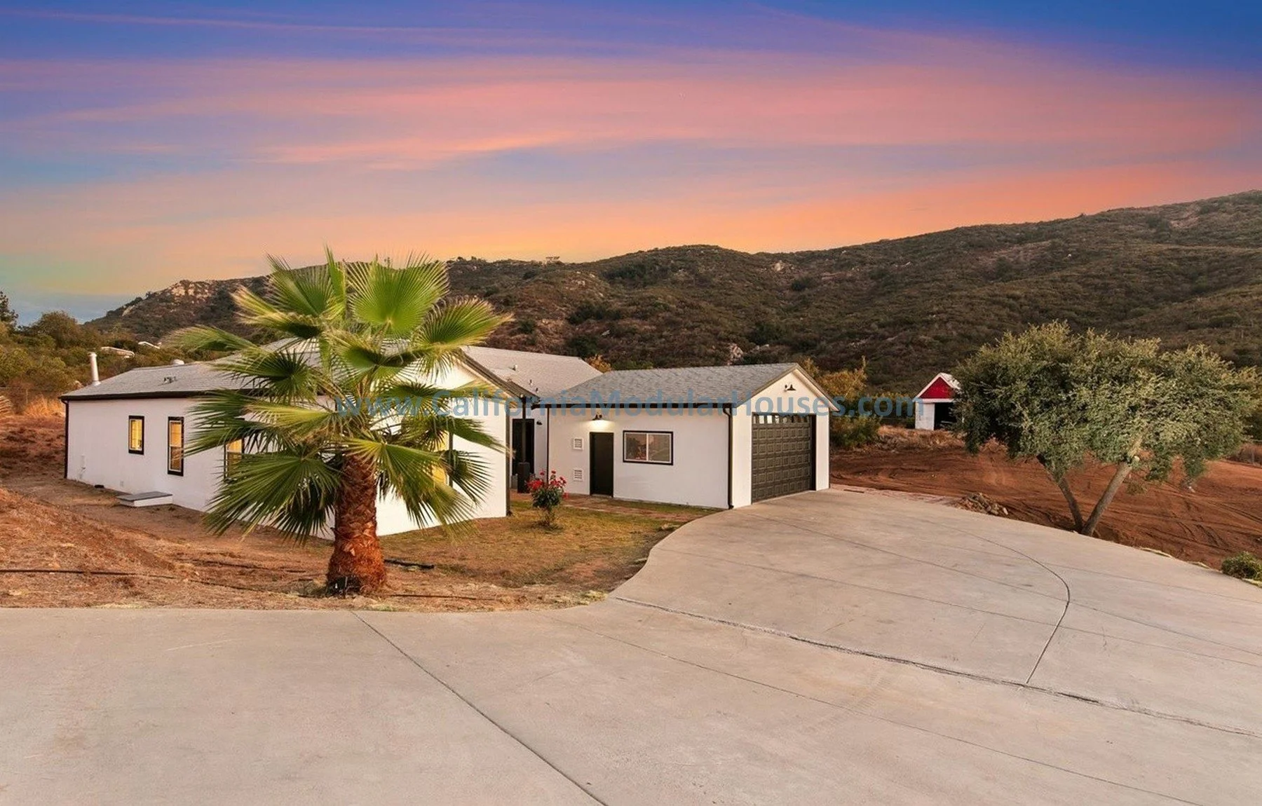 Modern white house with black accents and garage, palm tree and large tree in front, dirt and hillside landscape, colorful sunset sky in California.