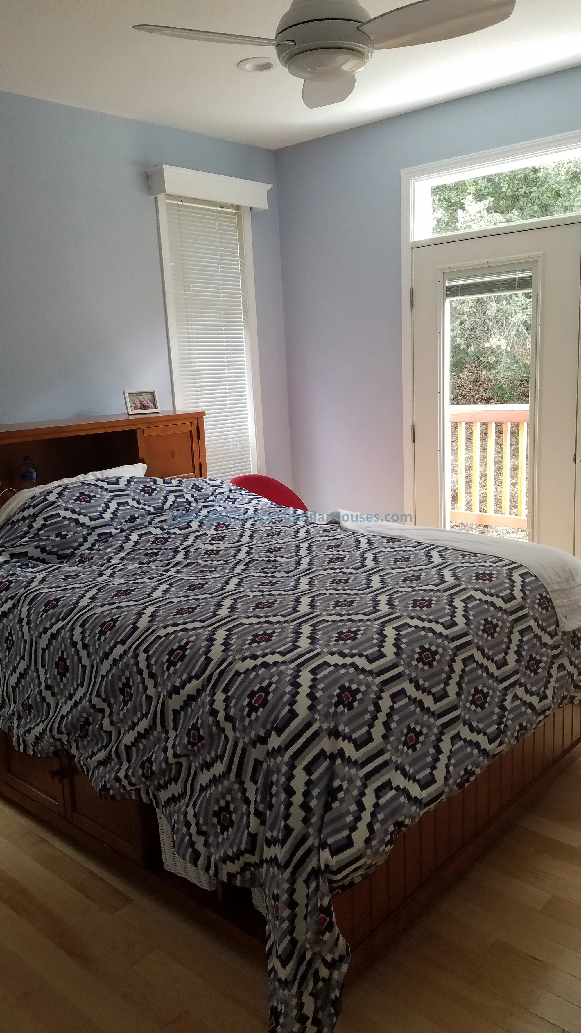 Bedroom with a wooden bed frame, patterned black and white bedspread, white walls, a window with blinds, a French door leading to a balcony, a ceiling fan, and a small framed photo on the nightstand.