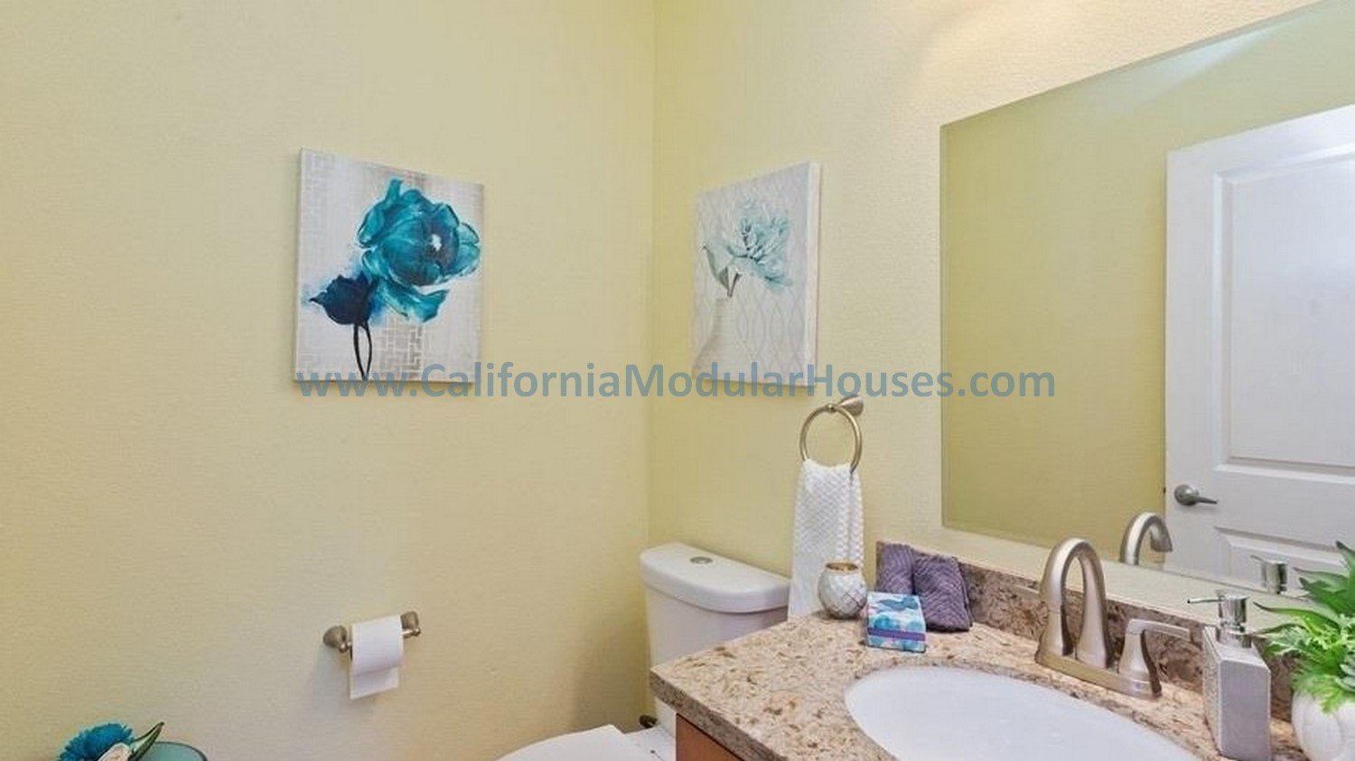 A bathroom with light yellow walls, two blue flower paintings, a granite countertop with a sink, a large mirror, a white toilet, and a towel ring with a white towel.  Modular homes for California