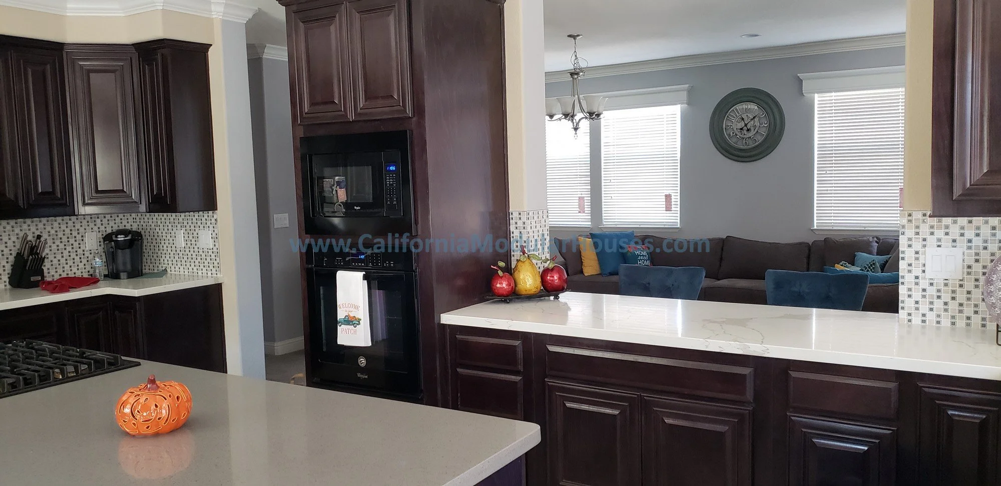 A modern kitchen with dark wooden cabinets, a white marble countertops, with a view into a living room to make sure that everybody is included, the people at the barstool area, the living room, and the kitchen. Pre-Fab.  