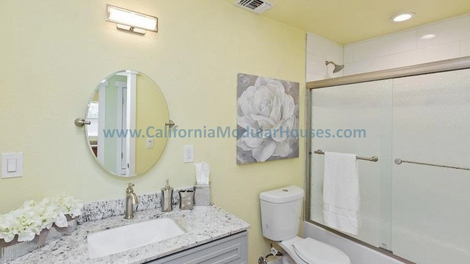 Bathroom with yellow walls, oval mirror, granite countertop with sink, flowers, tissue box, artwork of a white flower, toilet, glass shower with towel bar, ceiling light, and vent.  San Francisco Bay Area, CA California modular home.  prefab.