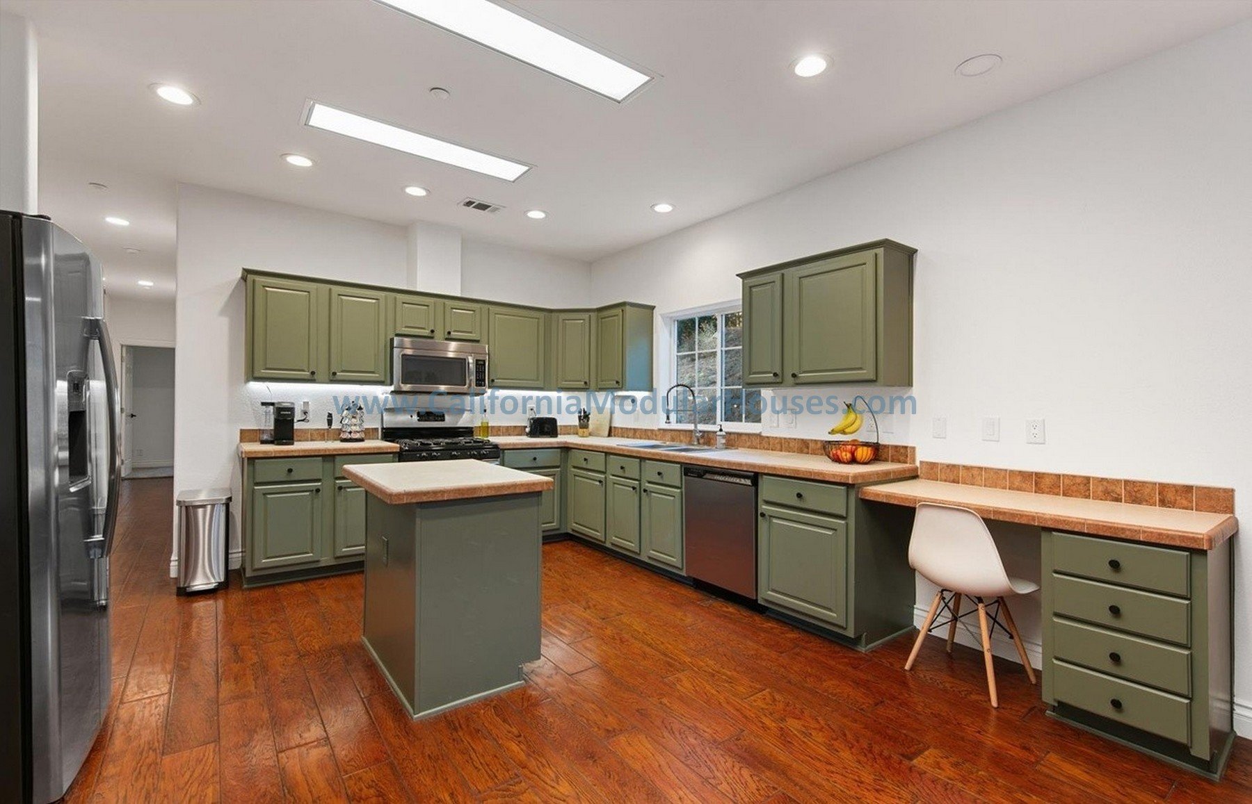 Modern kitchen with green cabinets, wooden countertops, a small island, stainless steel appliances including a refrigerator, microwave, and dishwasher, a window above the sink, and a white chair at the corner.