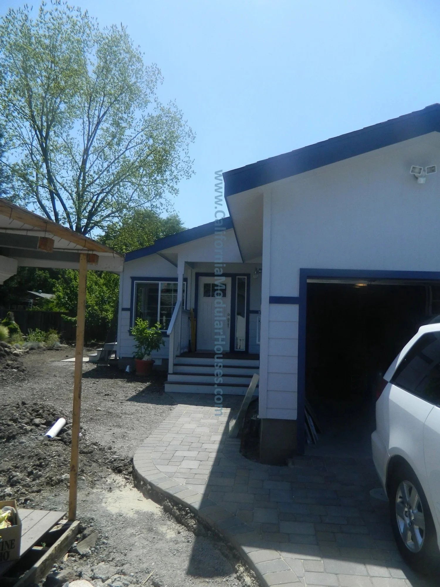 Newly constructed white house with blue trim and garage, front porch with steps, and a paved walkway. There's a car partially visible in the driveway, and a tree with green leaves in the background.