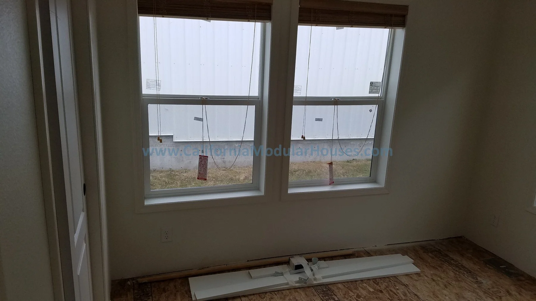Interior view of a room with two large windows, partially installed blinds, and construction debris on the wooden floor. Outside the windows, a white wall is visible.  Two-Story Modular Home. Pre-Fabricated Homes.