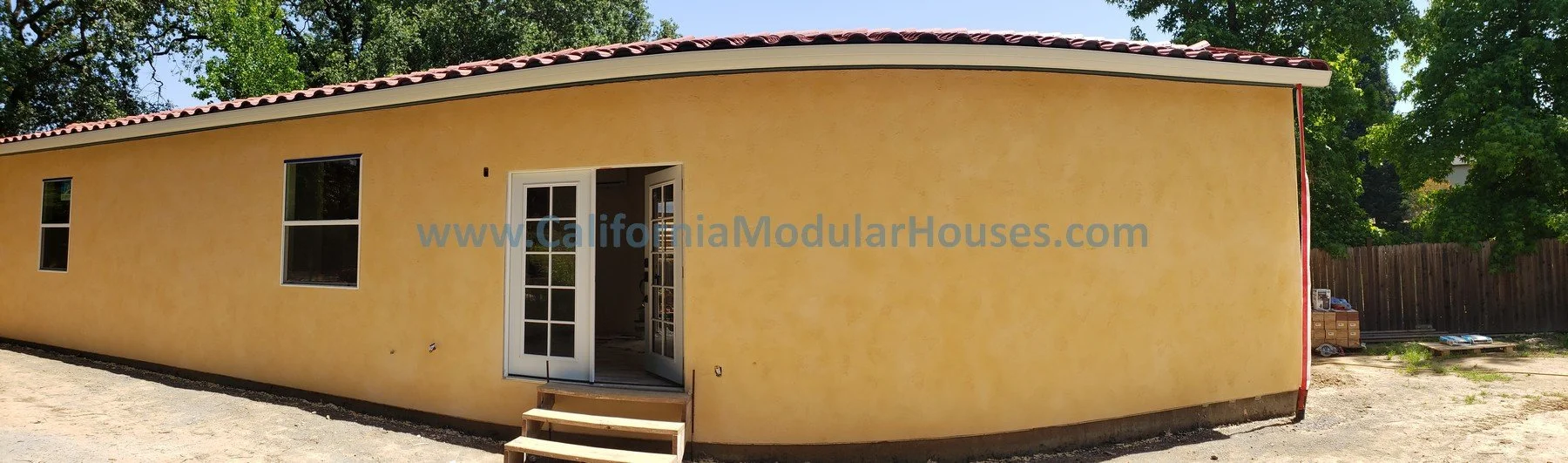 Yellow modular house with a red-tiled roof, two windows, and a glass door with steps leading up to it. Construction materials are on the ground nearby, and there is a wooden fence and trees in the background.  Healdsburg, Sonoma County, CA. 