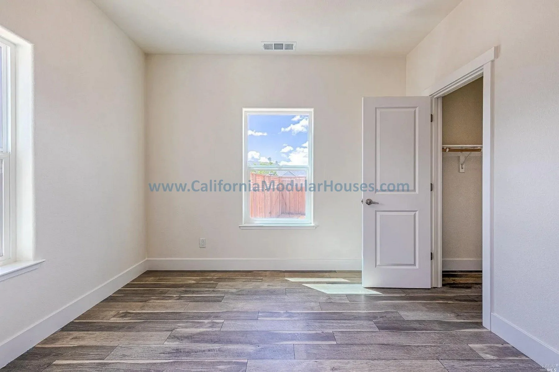 Empty room with white walls, wooden flooring, a window showing a fence and a cloudy sky, and an open closet door.